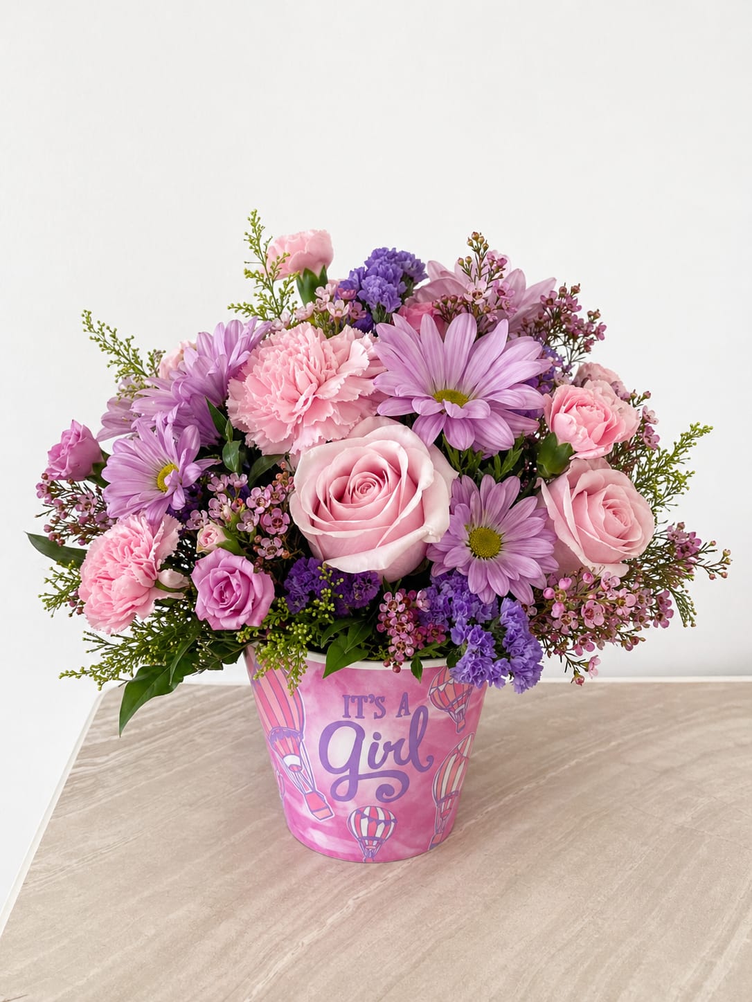 It’s a Girl Floral Arrangement - Sweet, delicate, and full of charm, this arrangement is the perfect way to celebrate a new baby girl. Designed in an adorable “It’s a Girl” keepsake container, it features a beautiful blend of soft pink roses, blush carnations, lavender daisies, and dainty filler flowers, all accented with fresh greenery.  The mix of pastel pinks and gentle purples creates a dreamy, feminine look that feels both joyful and elegant. Perfect for baby showers, hospital deliveries, or welcoming a little one home, this arrangement adds a heartfelt touch to any special moment.  Handcrafted with care, each design is uniquely arranged to create a full, balanced, and beautifully styled piece that will be remembered long after the flowers are gone.