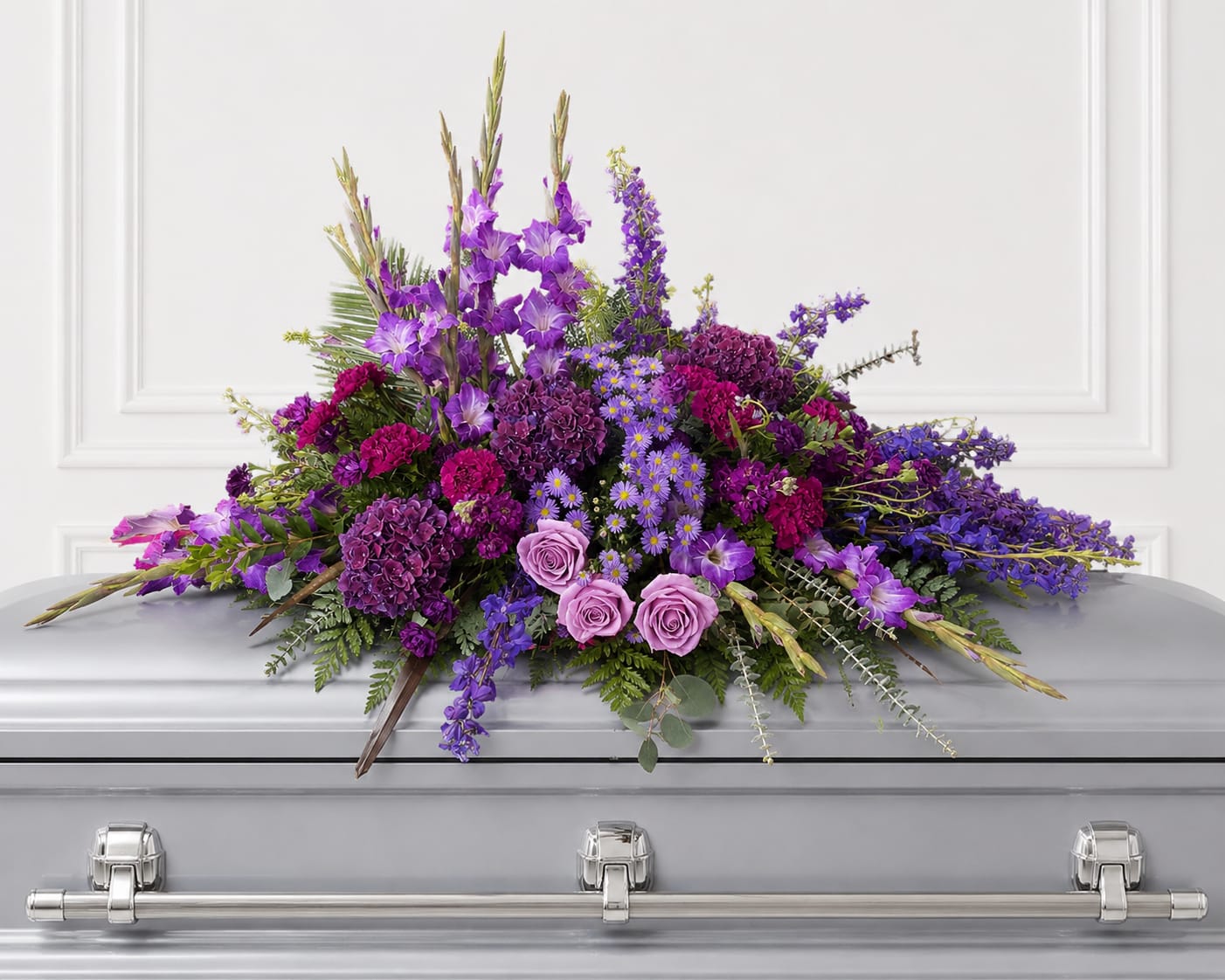 Golden Hour Casket Spray - A dramatic sweep of purple, magenta and lavender sets the tone for this casket spray, designed to offer a heartfelt farewell at a funeral or memorial service. The Golden Hour Casket Spray is a formal sympathy arrangement that reads as both ceremonial and deeply personal-the pale pink roses near the front add a gentle, comforting focal point while the overall palette honors the solemn occasion.  This large spray combines layered textures for a full, cascading silhouette: rounded roses and carnations, small daisy-like asters, and taller spiked accents that create vertical movement and graceful drape over the casket. Lush greenery frames the blooms and softens the composition, giving depth and a natural flow from front to back. The mix of purples and green ensures the tribute feels rich without overpowering the setting.  Made to rest on a standard casket, this arrangement is intended to convey deep sympathy and respect-an expression that will help the person you're thinking of feel remembered and honored. 