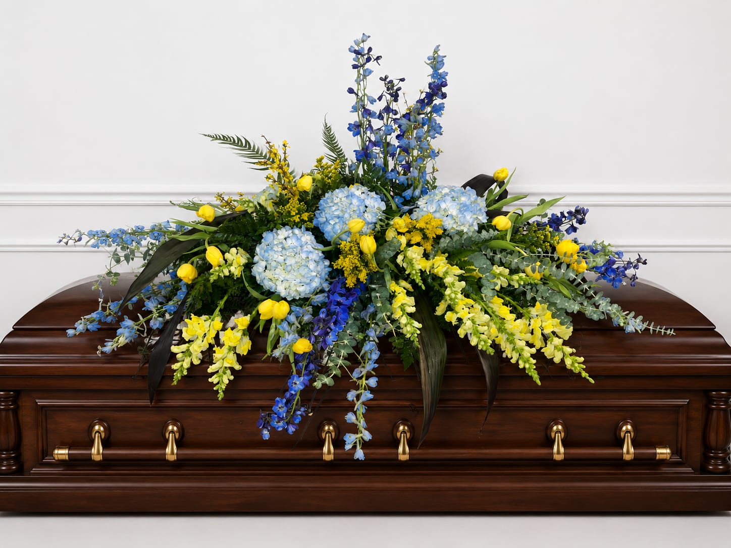 Timeless Essence Casket Spray - Pale blue hydrangeas and tall delphinium create a serene, comforting presence in this large casket spray designed for a funeral or memorial service. The blue-and-yellow palette - soft blue blooms balanced with sunny yellow snapdragons and petite yellow tulips - gives the arrangement a restrained brightness that reads respectful and uplifting for the person you're honoring.  Laid across a polished wooden casket, the broad, cascading shape delivers graceful coverage with dark foliage providing depth and contrast. Textures range from the rounded hydrangea heads to the vertical delphinium and snapdragon spikes, offering visual movement while keeping a formal silhouette appropriate for a solemn setting.  For families seeking a meaningful tribute, this spray communicates calm dignity and gentle warmth; it's the kind of arrangement that helps mourners feel remembered and comforted. 
