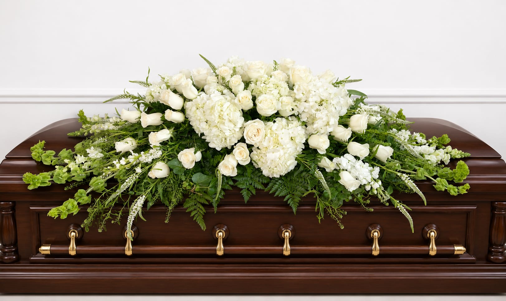 Heavenly Casket Spray - A large, all-white casket spray that reads as quietly majestic-Heavenly Casket Spray offers a serene tribute for a funeral service or memorial. This funeral flower arrangement combines white roses, lush white hydrangeas, and graceful stock to create a dignified statement piece that honors the person you've lost and comforts those who gather to remember them.  Clustered blooms form a full, cascading silhouette across the dark polished casket, softened by trailing greenery that adds texture and gentle movement. The white-and-green palette keeps the look traditional and restrained while the mix of rose heads, hydrangea clusters, and tall stock provides a varied, tactile composition that reads beautifully from every angle.  Designed for memorial services and bedside goodbyes, this spray communicates sympathy and respect in a timeless way-perfect for family members who want a meaningful, elegant tribute. 
