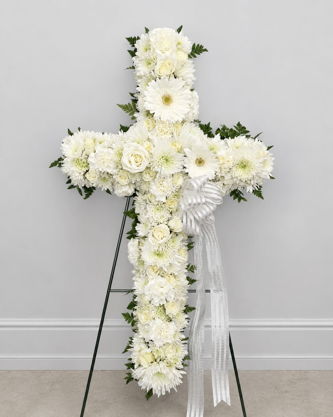 Sympathy Cross Standing Spray - A serene cross composed entirely of white blooms offers a quiet, reverent tribute for a sympathy or funeral service. The Sympathy Cross is arranged in the classic cruciform shape so the person you're honoring will be remembered with a dignified floral statement that reads from every angle.  Covered in white roses, chrysanthemums, and daisy-like gerbera blooms with soft touches of green foliage, the arrangement creates a layered, textured surface that feels both gentle and complete. A sheer white ribbon drapes gracefully from the right side and the piece is displayed on a metal easel for easy placement at a Funeral home, chapel, or graveside. The palette and form keep the focus on remembrance while providing a peaceful visual presence.  For families seeking a respectful way to express condolences, this cross conveys sympathy and support without words. 