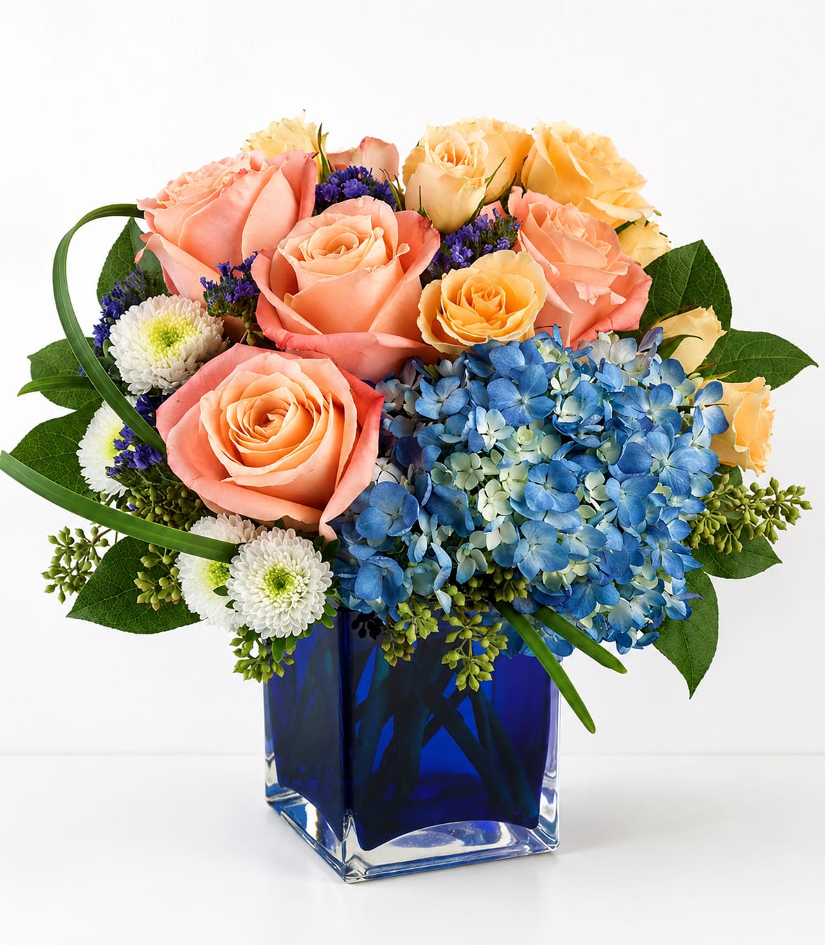 Pop of Color Blue Cube - A cheerful, modern bouquet that pops with color - peach roses and a vibrant cluster of blue hydrangeas create a fresh, celebratory feel for a birthday bouquet, thank-you gift, or springtime surprise. The strong contrast between warm peach and vivid blue reads joyful and uplifting, while the small white chrysanthemum accents soften the look so the recipient feels genuinely celebrated and seen.  This compact, rounded arrangement is presented in a deep cobalt-blue square glass vase that gives the design a polished, contemporary edge. Peach roses provide soft texture, the large blue hydrangea cluster offers bold volume, and the white mums add delicate highlights - a balanced mix that looks great on a dining table, desk, or entry console. Ideal for sending warm wishes or brightening someone's day; fits well in smaller spaces and arrives ready to display.