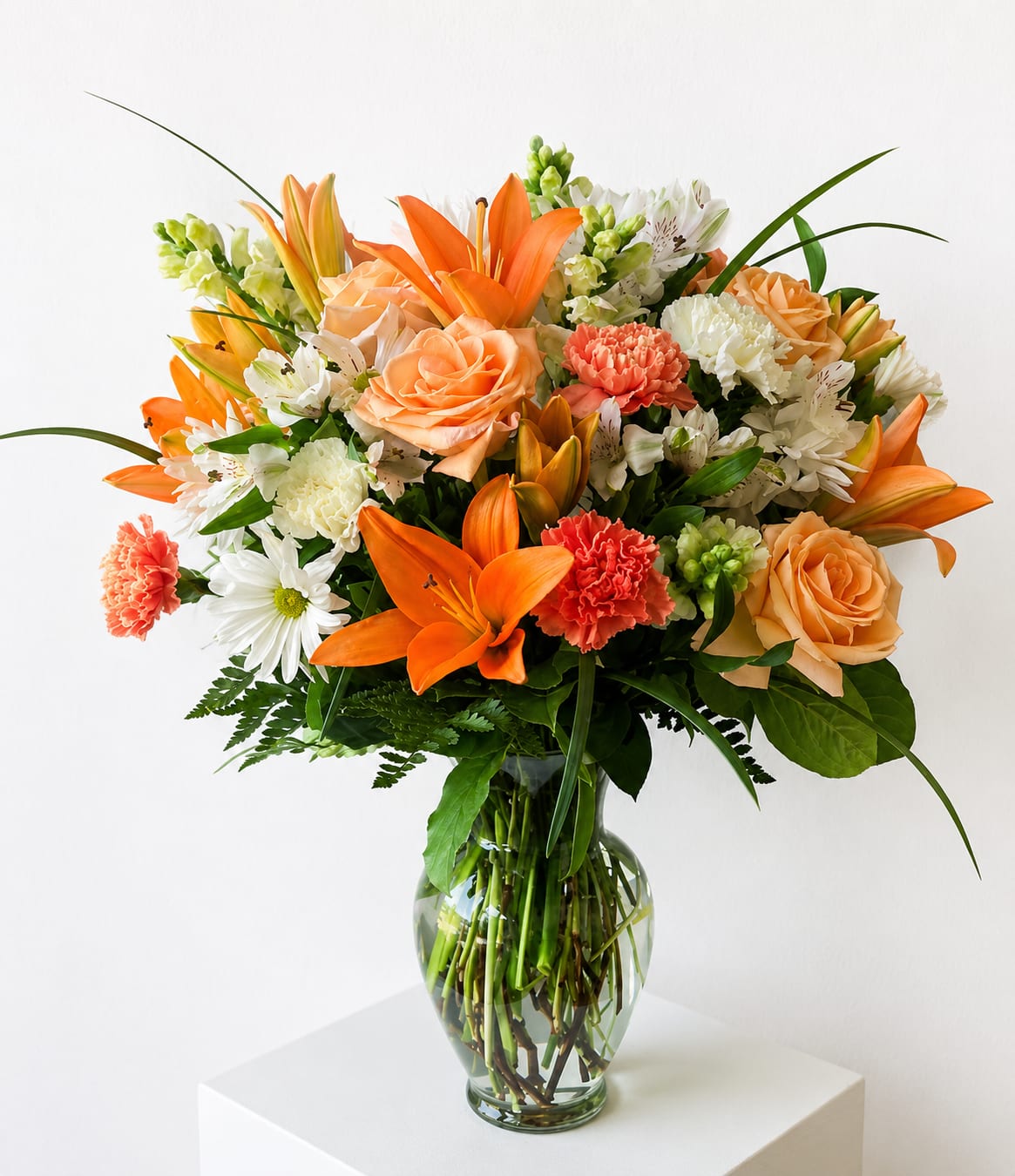 Golden Ray - A radiant mix of orange lilies and peach roses gives this flower arrangement a sunlit, celebratory presence-perfect as a birthday bouquet, congratulations gift, or a bright pick-me-up for someone you want to lift up. The warm orange and peach tones are balanced by white daisies and alstroemeria, so the recipient will feel both energized and genuinely seen when these cheerful blooms arrive.  This rounded design combines lilies, roses, carnations, alstroemeria, and daisies arranged in a clear glass vase, with long, arching blades and lush green foliage for a full, abundant silhouette. The coral carnations add textured warmth while the white blooms keep the arrangement airy and fresh. Sized to sit beautifully on a table or desk, it arrives ready to display-no extra fuss, just lasting color and a joyful impression.