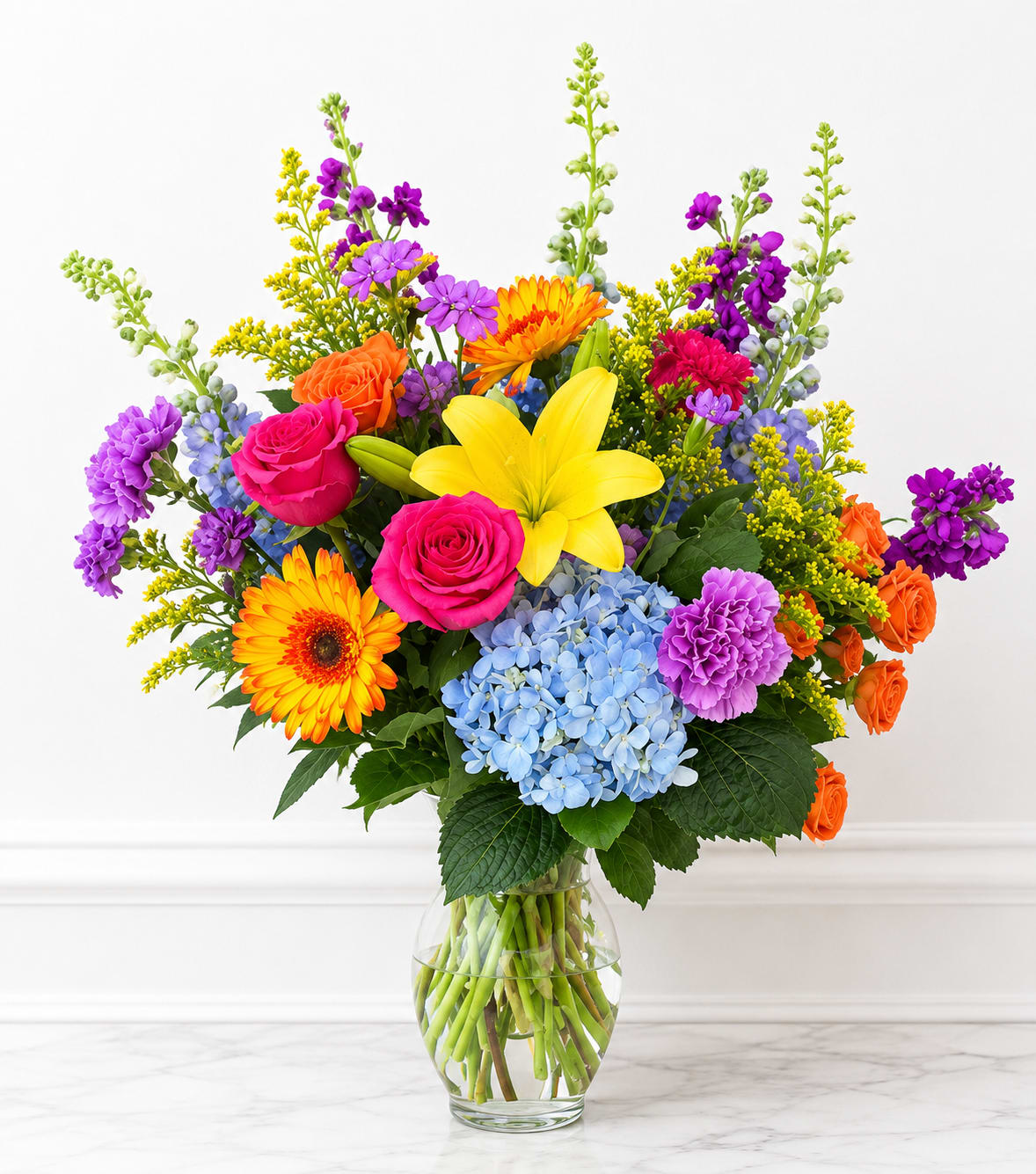 Beaming Bright Floral - A burst of warm color greets the recipient with Radiant Sunshine, a mixed-color flower arrangement perfect for birthdays, congratulations, or just because you want someone to feel celebrated. This bright, energetic bouquet reads like a party in bloom-the sort of gift that makes the person you're thinking of feel noticed and genuinely cheered.  Packed into a clear glass vase, the design features hot pink and orange roses, a large yellow lily, blue hydrangea, orange gerbera daisies, purple accent flowers, chrysanthemums, snapdragons, and additional seasonal blooms for texture and movement. The arrangement is dense and upright with a full, abundant shape; strong contrasts between the vivid pinks, oranges, yellows, blues, and purples create lively visual impact and a joyful palette that photographs beautifully.  Ideal for marking special moments from milestone birthdays to big congratulations, Radiant Sunshine is the kind of bouquet that brightens a room and uplifts the recipient. Vased and ready to display, the arrangement arrives assembled-refresh stems in water on arrival and keep in a cool spot out of direct sun to extend freshness.
