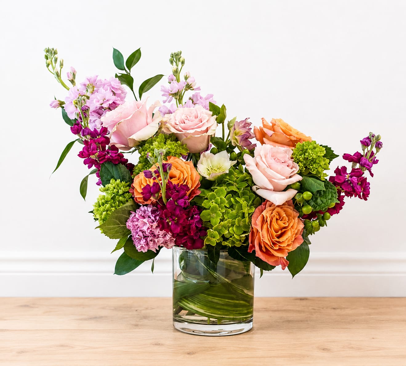 Springtime Blooms - Pale pink and peach roses meet bright orange blooms and vivid green hydrangea-like clusters in a lively, garden-style bouquet that reads as both celebratory and romantic - ideal as a birthday bouquet, anniversary arrangement, spring gifting, or a thank-you surprise. The mixed-color palette and rounded, full shape give the recipient an immediate sense of warmth and joy; she'll feel noticed and celebrated when these cheerful colors arrive at her door.  This flower arrangement combines roses and hydrangea-like accents with delicate stock and additional mixed blooms for texture and movement. Flowers fan outward from a clear cylindrical glass vase to create a generous, hand-tied look that's easy to display on a dining table or entry console. The blend of pinks, peaches, orange and green offers visual contrast and soft fragrance without overwhelming the room.  Perfect for sending to someone who loves fresh, garden-inspired style, this piece is a thoughtful way to mark a milestone or say "thank you" in color. Arrange on a steady surface and change vase water every 2-3 days, trimming stems slightly to extend freshness; order early for spring delivery to match seasonal availability.