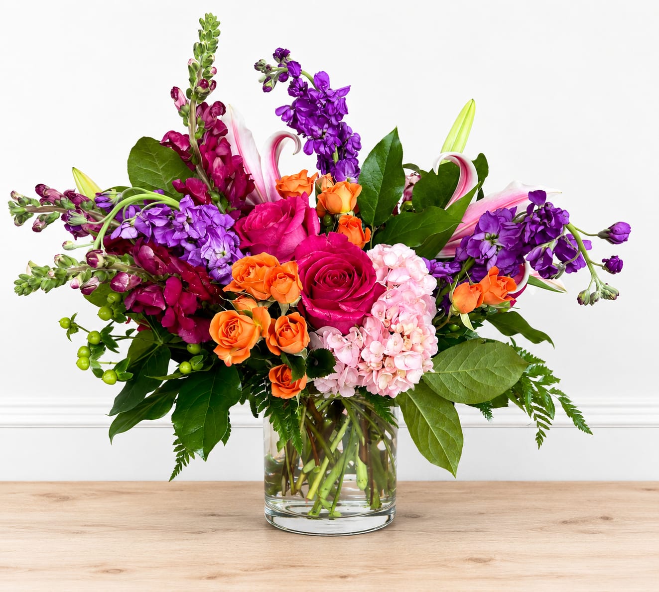 Violet - Vivid magenta roses and purple snapdragons create an immediate burst of color in this flower arrangement, making it a joyful choice for a birthday bouquet, congratulations, or just to brighten someone's day. The lively mix of purple, pink, orange and white reads festive and personal - the person you're thinking of will feel noticed and celebrated the moment these colors arrive.  A broad, airy composition pairs tall, architectural snapdragon spikes with arching pink-and-white lilies and clusters of soft pink hydrangea, while orange spray roses and saturated magenta roses add punch and texture. Arranged in a clear cylindrical glass vase, the asymmetrical design lets blooms extend outward for movement and presence on a table or desk. The varied heights and forms give this piece a fresh, garden-picked look without being fussy.  Perfect for cheerful gifting - birthdays, congratulations, or an everyday surprise - this arrangement is delivered ready to display in its clear glass vase. For best longevity, place away from direct heat and change the water every couple of days so the recipient can enjoy the bright colors and fragrances longer.