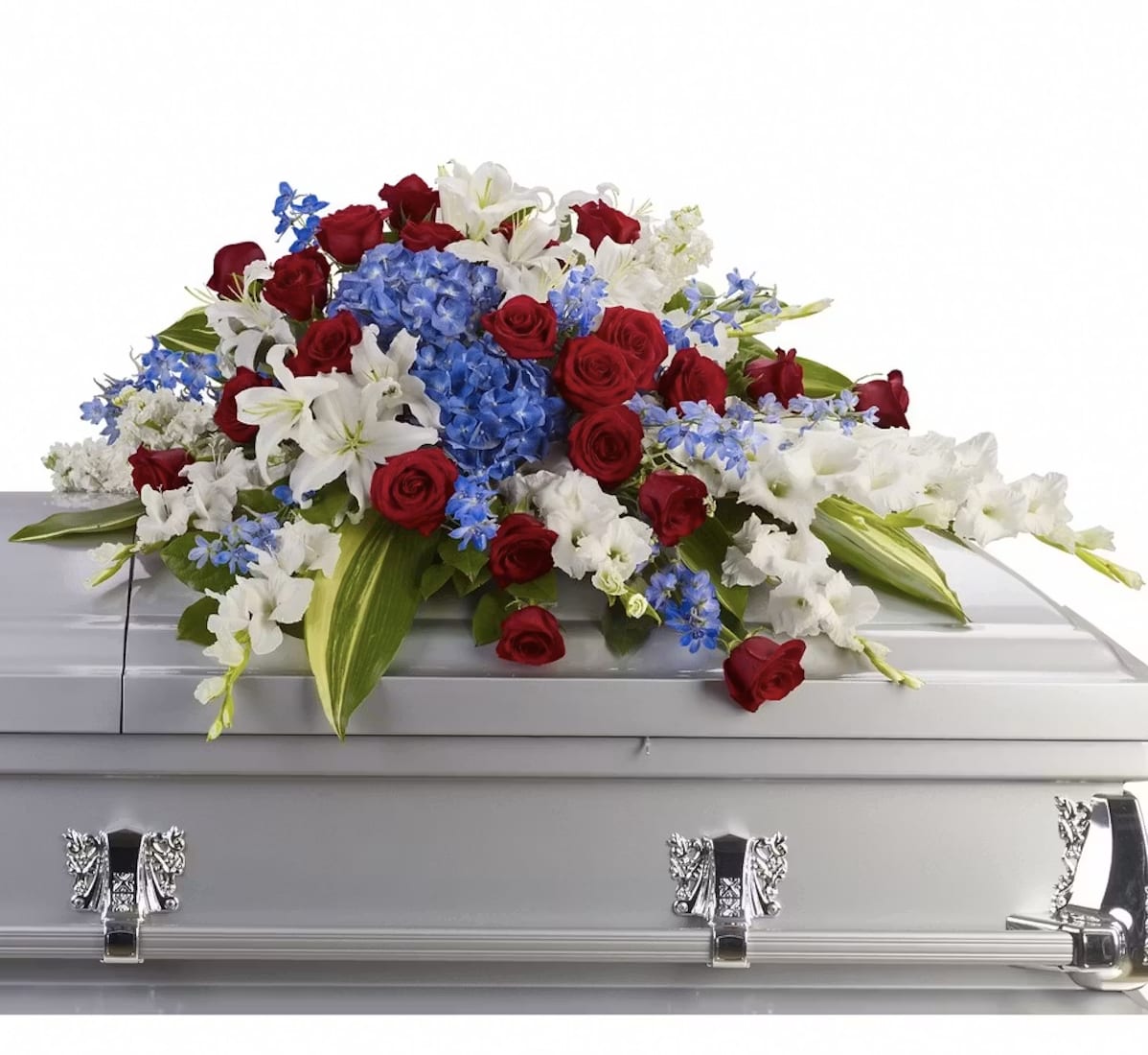 A Tribute to a Cherished Soul - A broad, low casket spray that lays like a final embrace, its dense cascade of red roses, white lilies and blue hydrangea arranged for a dignified funeral tribute. This flower arrangement is designed specifically as a casket piece for memorial services and patriotic remembrances, the red, white and blue palette creating a solemn, respectful tone while the white lilies lend gentle comfort.  A ceremonial style, blooms spill outward over the edges in a generous, cascading cluster. Textural contrast comes from red roses, the sculpted trumpet shape of lilies, full hydrangea heads and small blue filler flowers set against green foliage - a composition meant to read clearly from the front of the casket. For a meaningful farewell that honors their life and service, this spray arrives ready to place and is intended for use at memorials and funeral services.