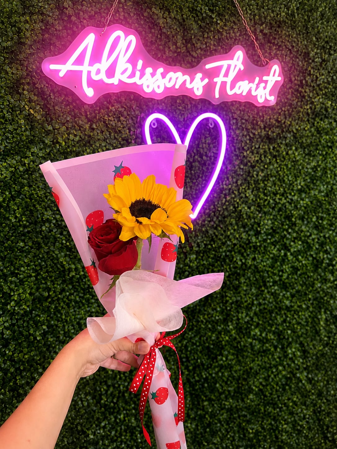 Strawberry Kiss Bouquet - Brighten someone's day with this charming hand-wrapped bouquet featuring a vibrant sunflower paired with a classic red rose. Wrapped in our adorable strawberry-themed paper and finished with a coordinating ribbon, it's the perfect little gift for birthdays, anniversaries, celebrations, or just because.   Please Note:  Pickup in store only – this item is not available for delivery. Prefer a different rose color or wrapping paper? We'd be happy to customize it! Simply call us to place your order, and we'll do our best to accommodate your request based on flower and paper availability.  A simple, thoughtful bouquet that makes a big impression. 