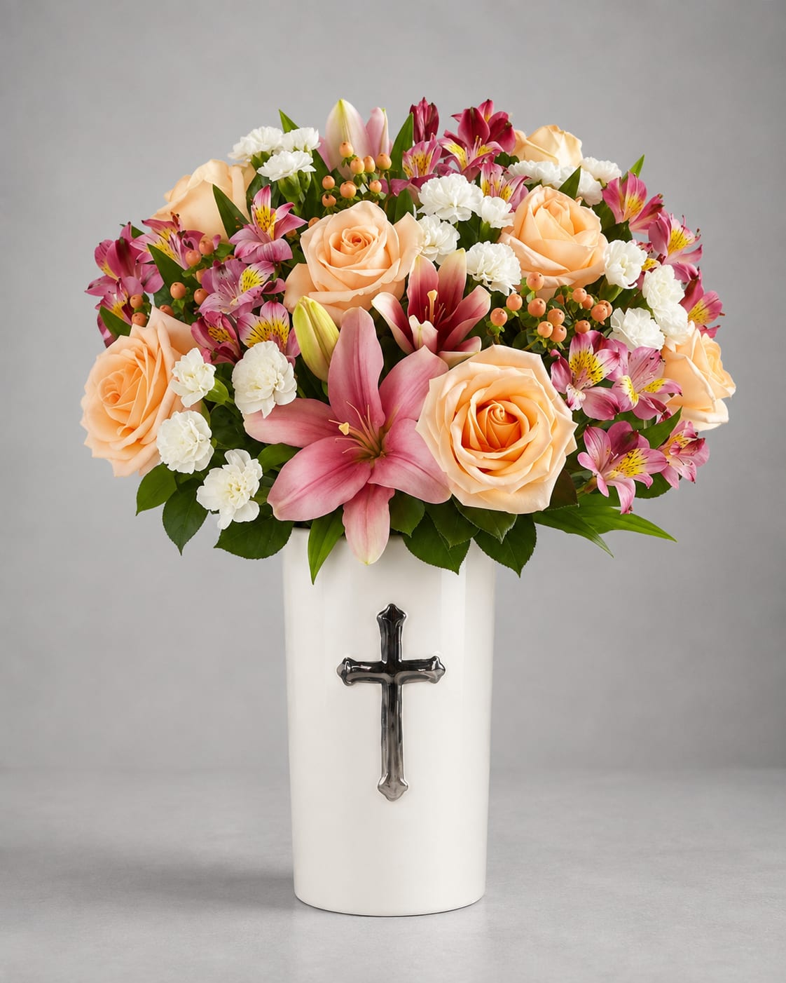 Peach roses and pink lilies in a white vase with a cross