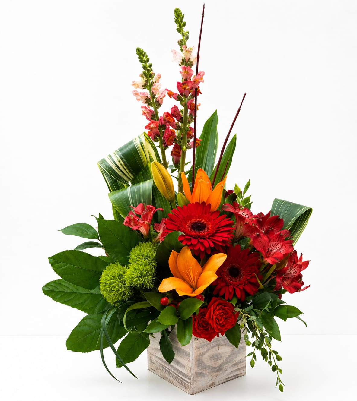 Wooden Box Floral Arrangement  - A compact wooden box overflowing with bold red roses, fiery orange lilies, and bright pink alstroemeria creates a glamorous birthday bouquet that reads as both romantic and celebratory. The arrangement's densely packed red roses, some with a subtle glittered finish, give a dramatic, gift-ready focal point while red gerbera daisies and green accent blooms add cheerful contrast-perfect for love and romance, a heartfelt thank you, or a "just because" surprise.  Built in a modern wooden cube vase, the arrangement uses layered black, white, and red decorative paper, a wide black ribbon that reads "HAPPY BIRTHDAY," and a mix of silver crown and butterfly toppers for an ornate, festive look. Broad glossy leaves, striped decorative leaf wrap, floral picks and a tasteful silver crown topper add texture and height without feeling fussy. The color mix of pink, red, orange, green, black and white gives a lively, energetic presence that suits birthdays, congratulations, or brightening someone's day.  Send this when you want the person you're thinking of to feel truly celebrated-romantic enough for an anniversary-style moment, playful enough for a milestone birthday, and confident as a thank-you gift. The wooden cube makes a stylish keepsake vessel; for best appearance, change the water every other day and trim stems if flowers are rearranged into a vase.