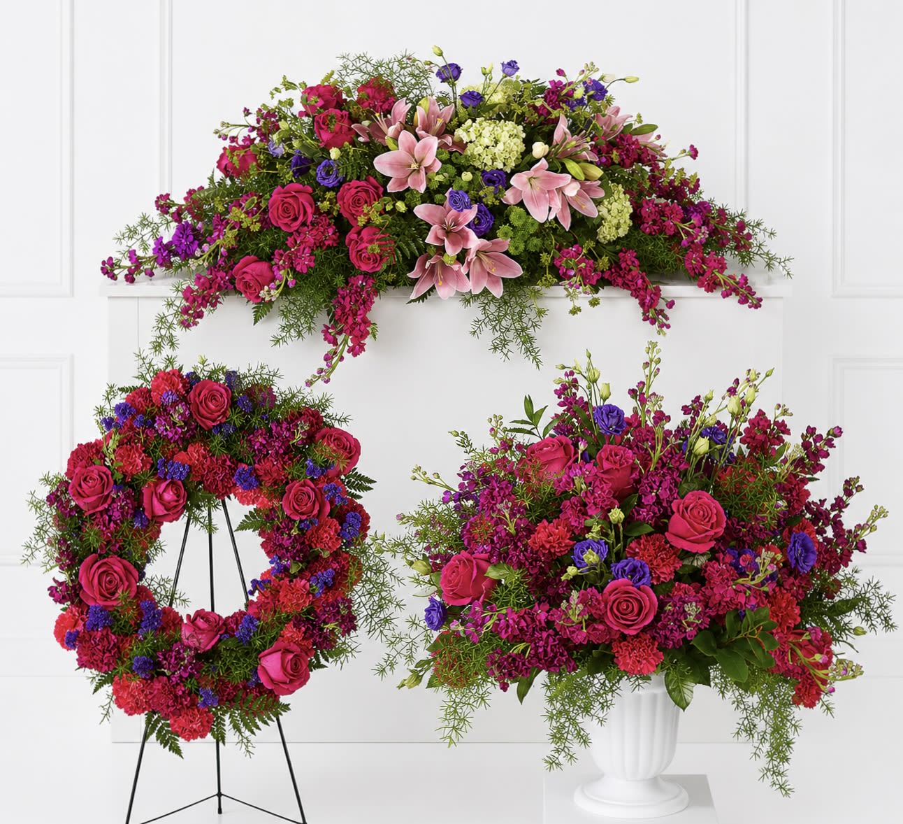 With Our Love Bundle  - A vivid, multi-piece floral bundle that reads like a comforting sympathy and funeral tribute depending on delivery and placement. This flower arrangement set pairs bright hot-pink roses and pink lilies with purple accents.  The bundle includes three large floral designs: a wide, low standing spray for tabletops, a wreath mounted on a black metal easel for formal memorial displays, and a full, rounded arrangement presented in a white pedestal vase. Blooms include roses, lilies, carnations, hydrangea-like clusters, stock and dianthus alongside silk orchids and assorted filler flowers .  