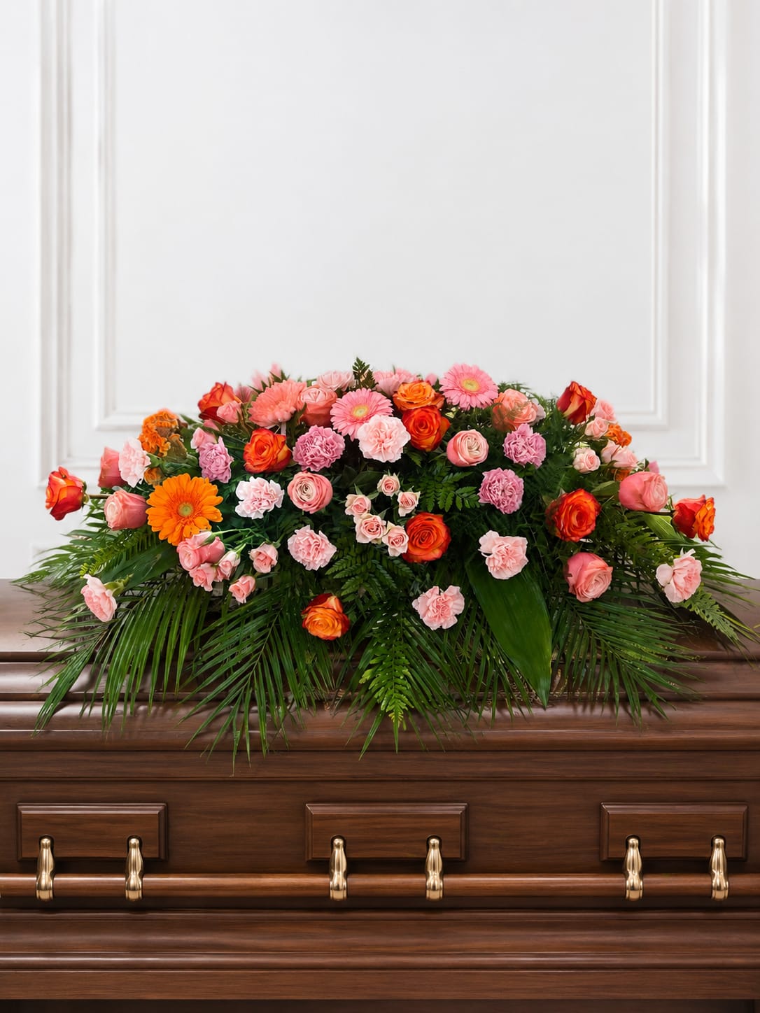 Graceful Casket Spray - A broad, rounded casket spray that brings a warm, expressive touch to a sympathy or funeral service, blending delicate orchids and classic roses with cheerful daisy-like blooms. The Graceful Casket Spray is arranged to rest across the top of the casket, creating an abundant, layered presentation that comforts and honors the person you're remembering.  Soft pink and orange roses and carnations mingle with gerbera-style blooms, set against dense green foliage and trailing palm fronds for texture and formality. The colors read bright yet respectful-white accents calm the palette while the greenery provides a full, structured backdrop that looks beautiful from every angle.  Designed for memorial services and to offer a meaningful tribute, this spray conveys warmth and remembrance for family and friends who gather to celebrate a life. Sized to span the casket with a graceful, rounded silhouette, it arrives wired and ready for placement by our experienced florist; please contact us for delivery timing and personalization options.