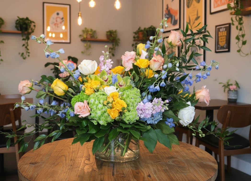 Ella's Garden - A lush, garden-style flower arrangement named after Ella, one of the talented floral designers here at Narcissus Florals, Ella's Garden Party is thoughtfully created for a lower styled aesthetic. This large, abundant mixed bouquet in a clear glass vase reads like a sunlit spring morning-perfect as a birthday bouquet, a just-because surprise, or a cheerful thank-you gift that makes the recipient feel truly celebrated.  Soft pastel pink and white roses mingle with bright yellow blooms, sky-blue accents, and a prominent green hydrangea cluster at the center for a fresh, balanced focal point. The design spreads outward in a loose, garden-style shape with long stems and airy branching elements, giving it a full, slightly dramatic presence that still sits low and elegant in its vase. Ideal for spring and Mother's Day or any moment that calls for warm, uplifting color, Ella's Garden arrives ready to display in a clear glass vase; simple care-fresh water and a cool spot-keeps the blooms bright for days.