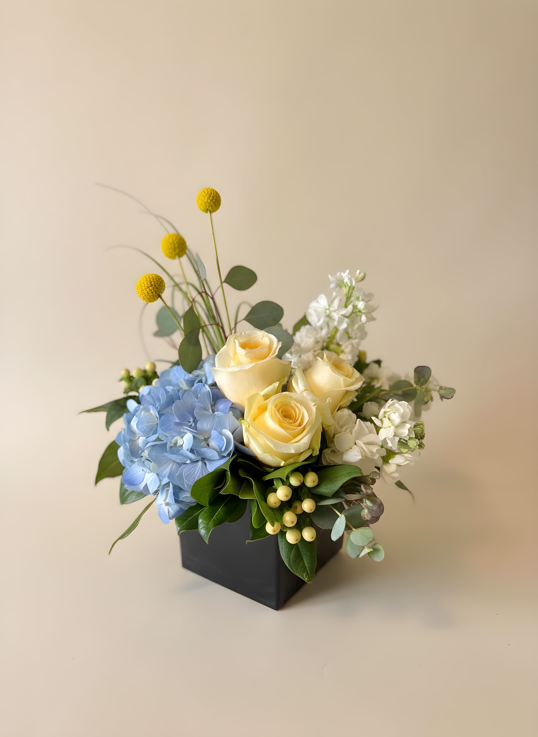 Blue Skies &amp; Buttercups - Pale blue hydrangeas and soft cream-to-yellow roses create an airy, calming sight in this compact modern arrangement-perfect as a cheerful flower arrangement that feels fresh and contemporary. Tall yellow billy balls punctuate the design, lifting the eye above the main cluster while white filler blooms and glossy green foliage add light, textural contrast against the dark vase.  Arranged in a sleek black square vase, the composition mixes hydrangeas, roses, stock, craspedia (billy balls) and other seasonal stems for a balanced, garden-fresh look. The bouquet is compact but full-bodied, with a gently rounded silhouette and upright accents that give it height without dominating a tabletop. Care tip: keep in fresh water, trim stems every few days, and place away from direct heat to prolong blooms.