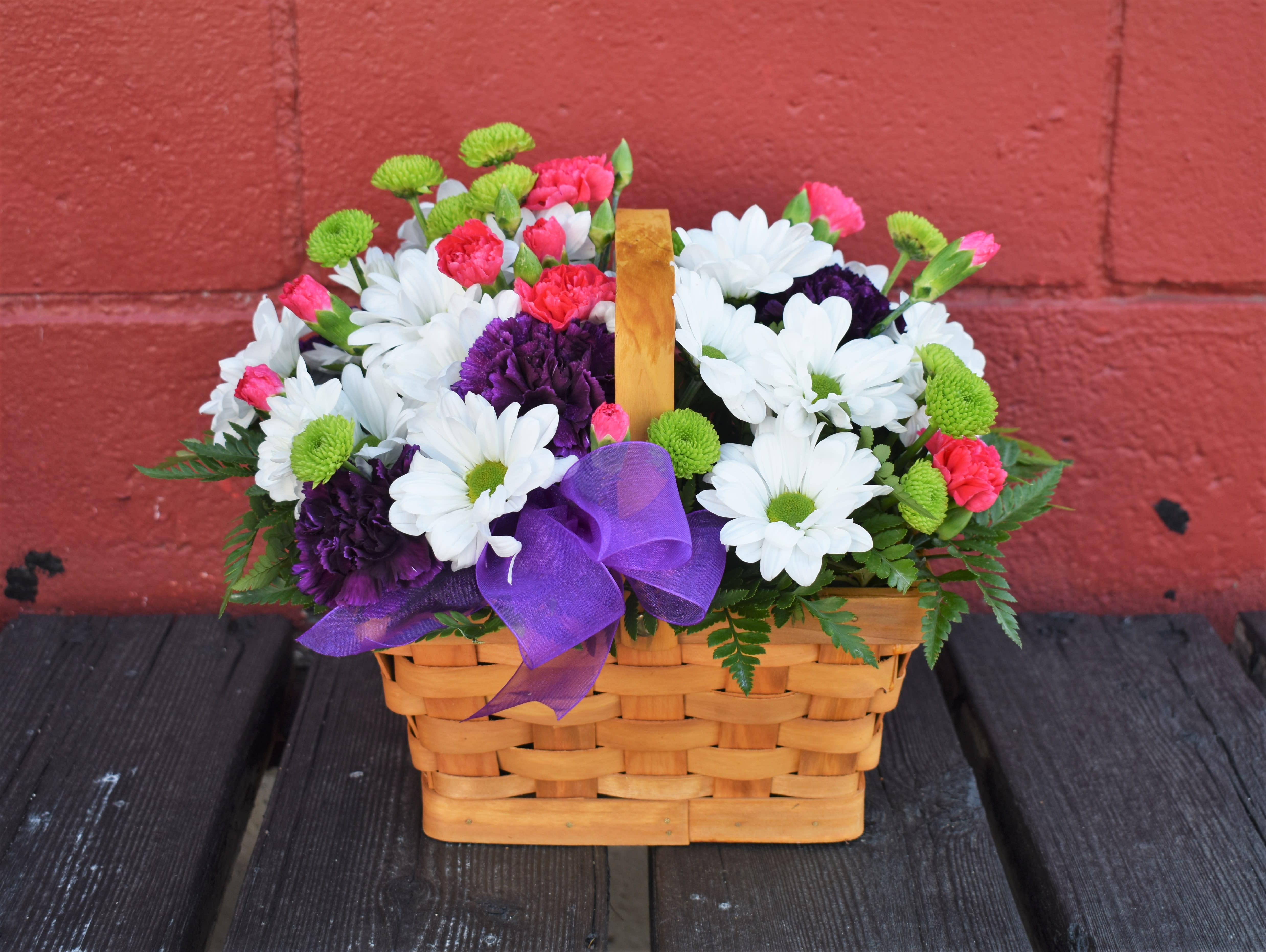 Blooming Bounty Basket - This pretty basket is sure to cheer up anyone's day. This handled basket is filled with green button pompons, white daisy pompons, hot pink mini carnations and purple carnations. A purple ribbon bow completes your best wishes.