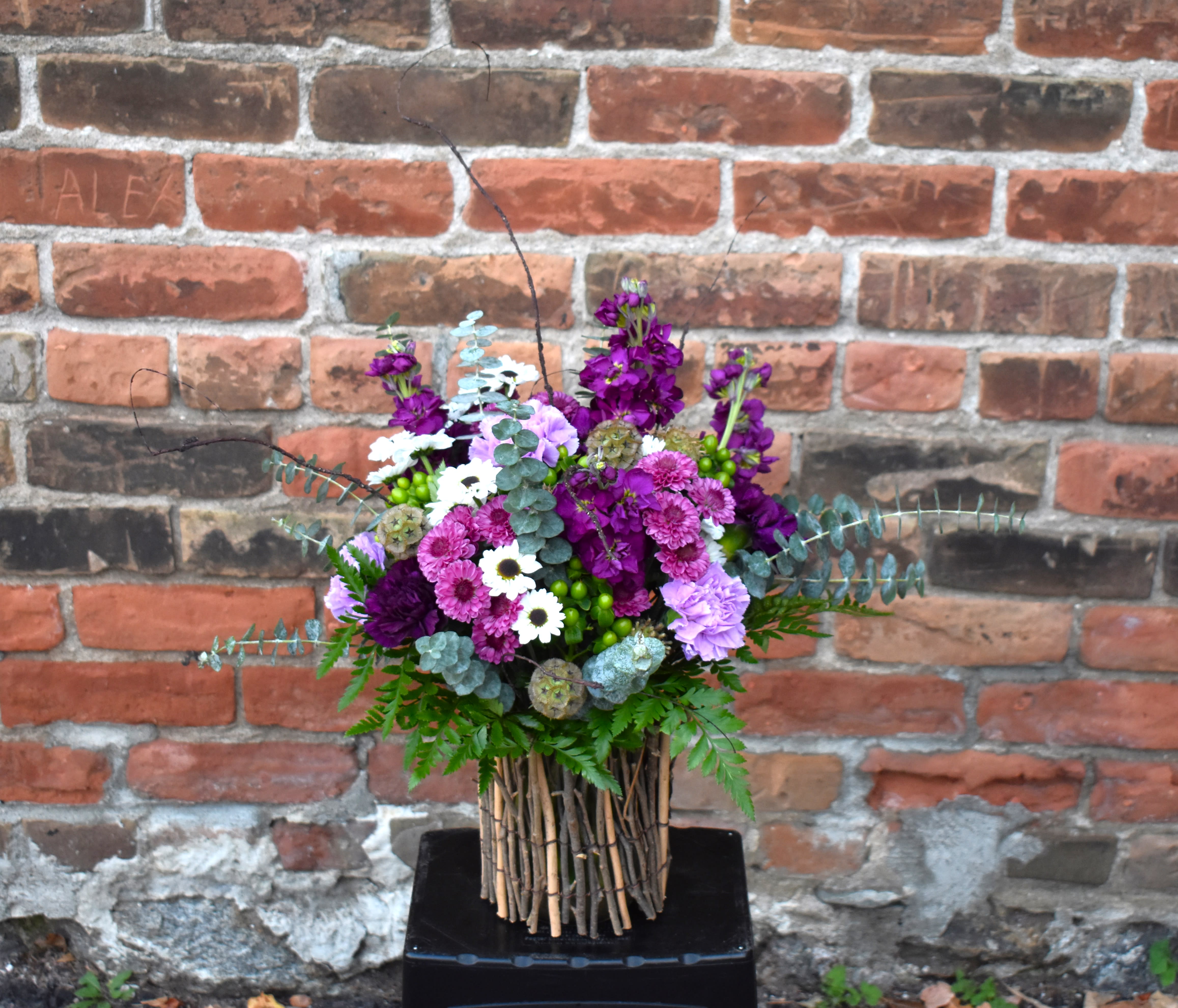Bewitched  - Rich purples and vibrant greens created with rustic twiggy elements combining for a bewitchingly magical bouquet! Purple stock, lavender and plum carnations, white and purple mums, green hypericum, and eucalyptus are designed in a twiggy vase and accented by rustic scaviosa pods...creating a naturally beautiful design!