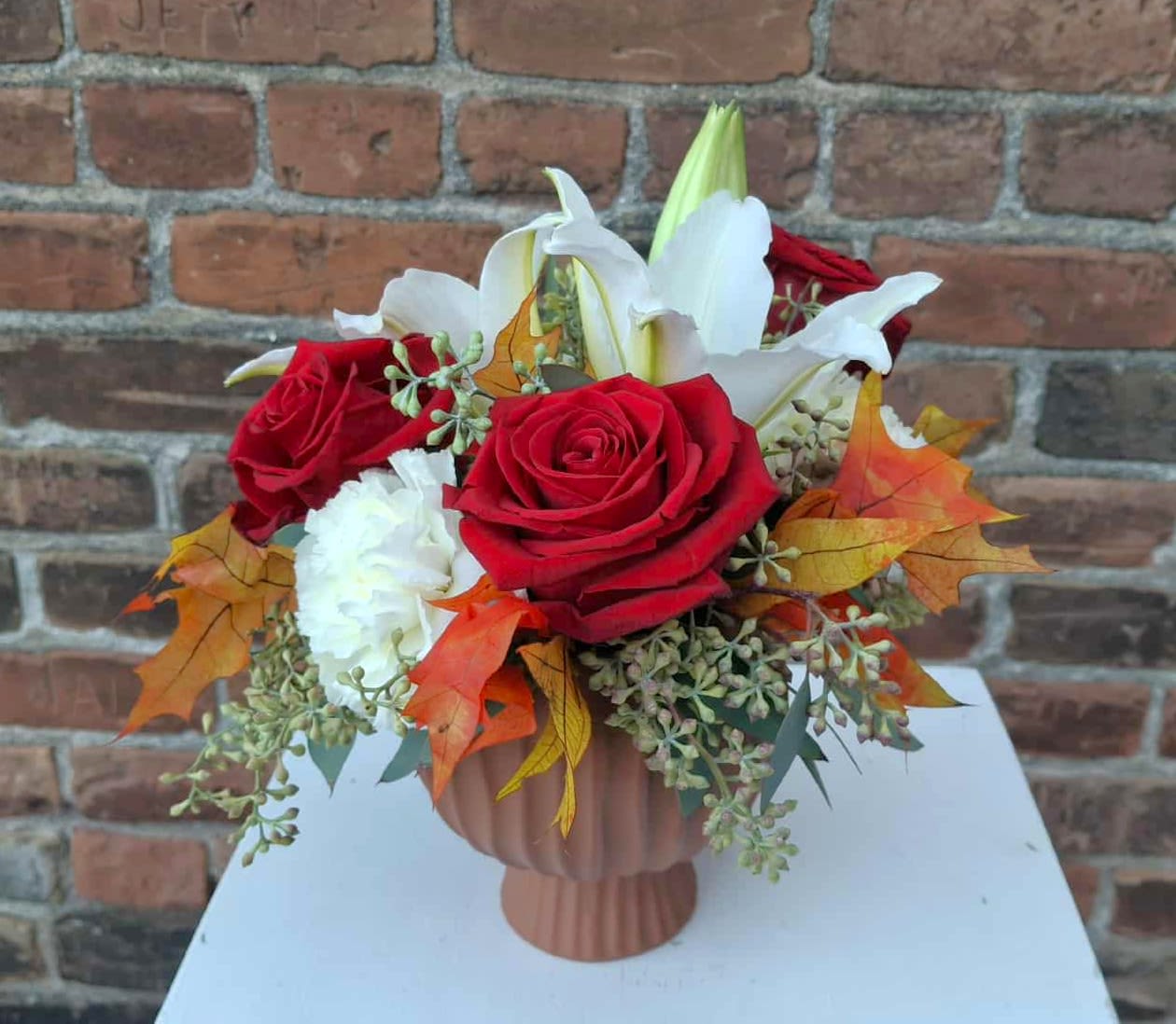 Creamy Harvest Hues - A vibrant and modern bouquet bursting with color and elegant blooms! red roses, white carnations, and white lilies are designed in terracotta pottery and accented with eucalyptus and fall leaves...the perfect way to celebrate the season!  Approx 8" X 8"