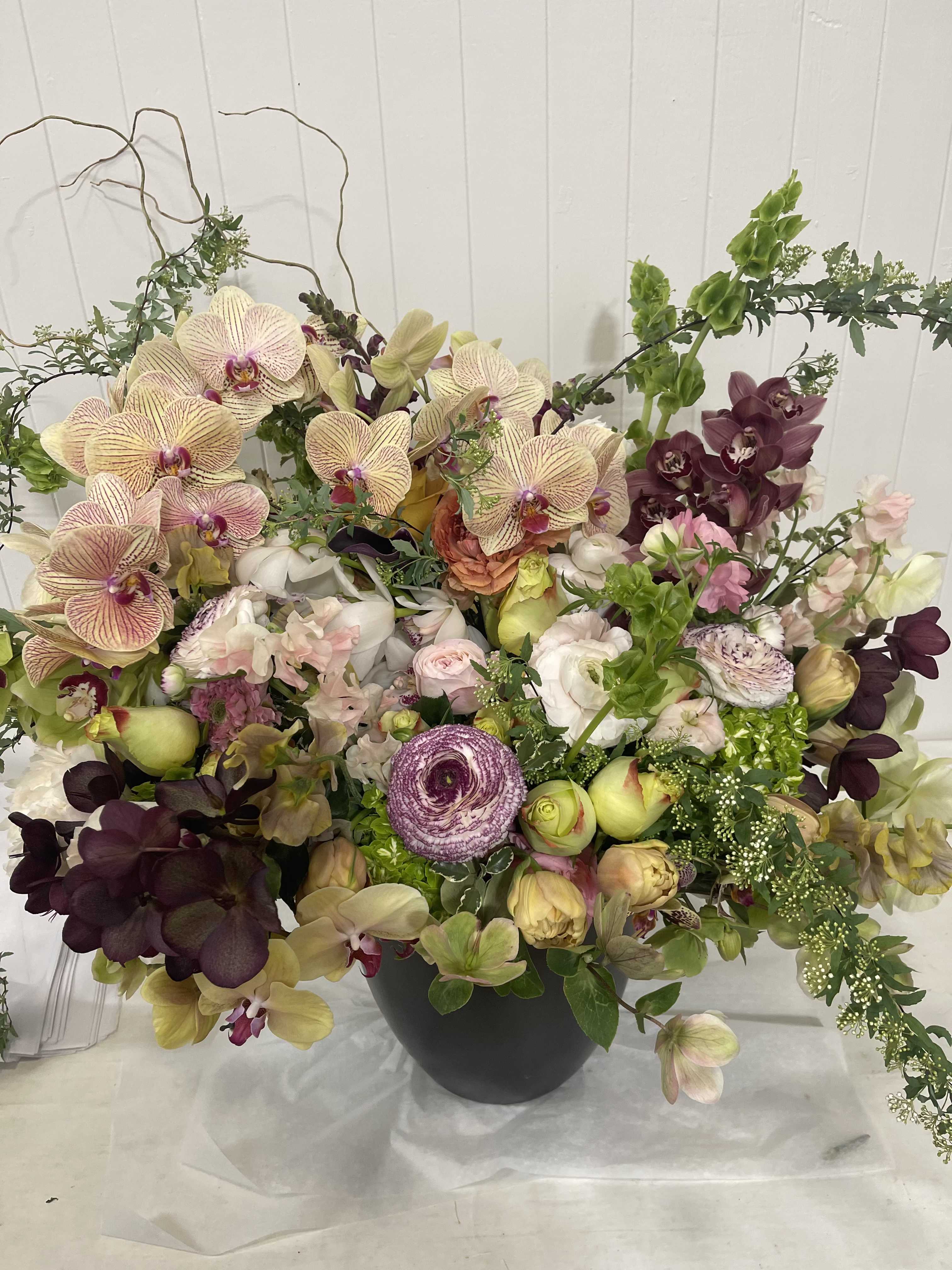 Large mixed orchid arrangement in a dark vase with pale and deep purple blooms