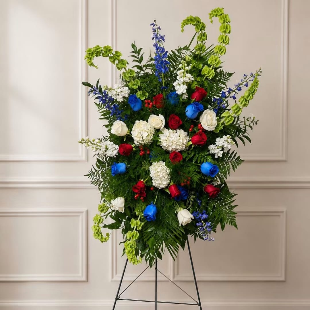 Our Hero Tribute Spray - A tall, ceremonial standing spray that commands attention with a patriotic palette-red, white and blue roses paired with white hydrangea-like blooms and sky-blue delphinium spikes-designed for memorials, Veterans Day, funerals, or any occasion honoring a hero. The arrangement's vertical silhouette and layered greenery create a respectful, formal impression so the person you're honoring will feel remembered and celebrated in a dignified way.  Built on a metal easel with a black easel stand, this large-scale spray is packed with abundant foliage and bright green hanging accents that arc above the composition for added movement and height. The dense, vertical form reads as a public display rather than a tabletop piece, combining classic roses and textured blooms for strong visual impact from a distance.  Ideal for commemorative services, patriotic ceremonies, or a prominent display at a memorial site, this tribute sends a clear message of honor and respect. Because it's mounted on an easel, it's ready to stand at the front of a service or beside a gravesite; no handheld support is needed.