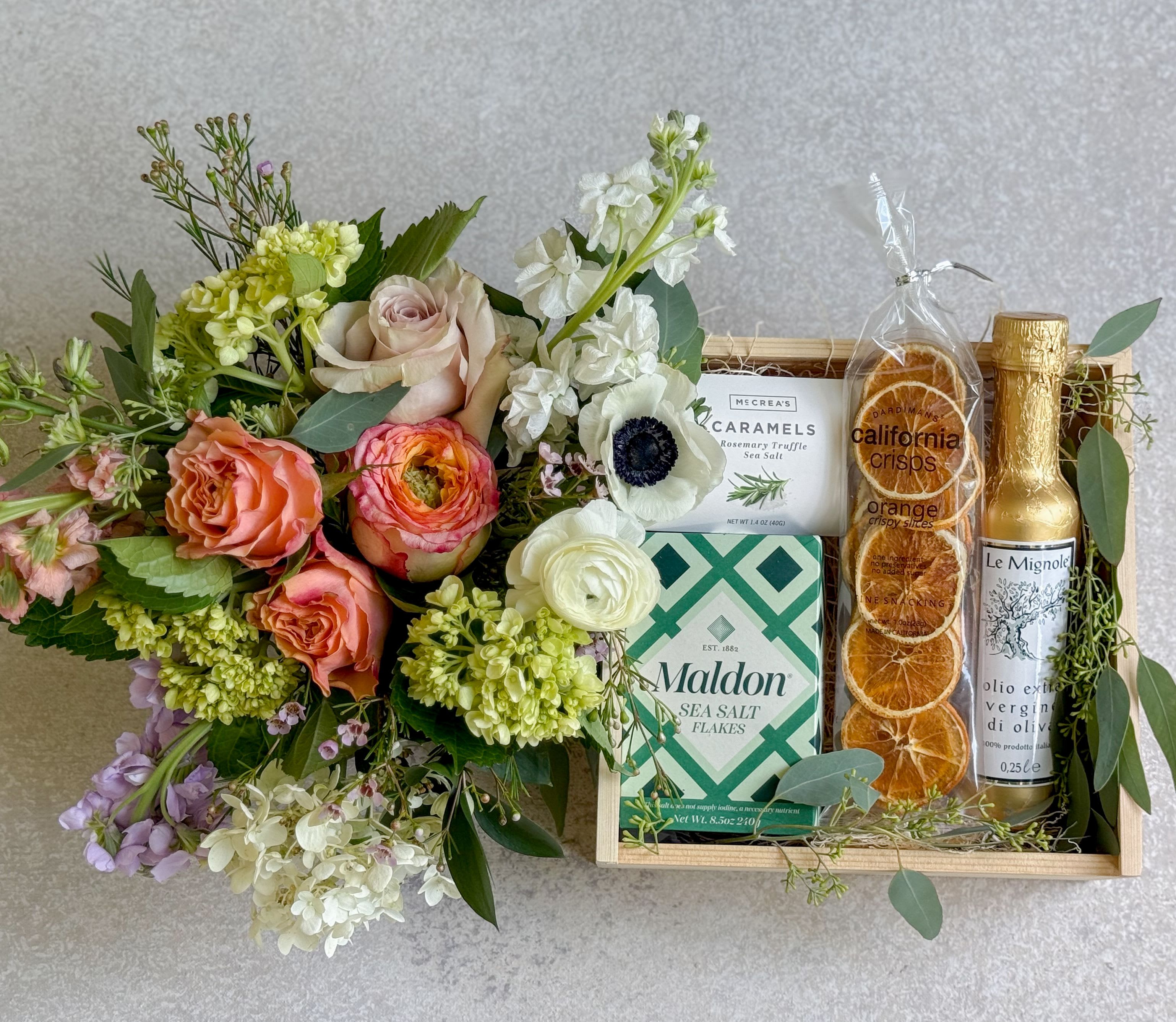 Floral bouquet beside a gift tray with caramels, sea salt, crisps, and olive oil