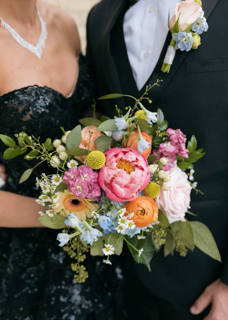 Colorful hand-tied bouquet of pink peonies, orange ranunculus, and mixed blooms held by a dressed-up couple