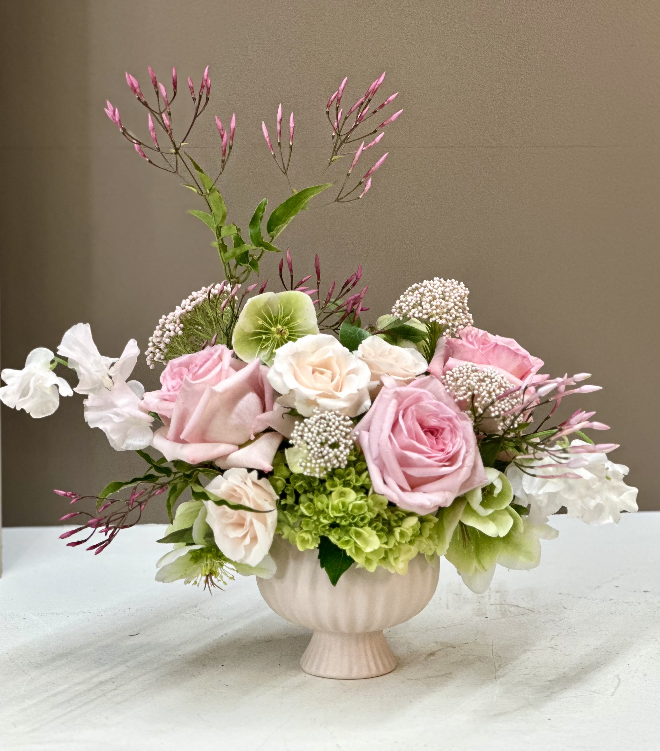 Blush Elegance - A soft blend of Blush Pink Roses, Hellebores, Sweet Peas, and airy Pink Jasmine, artfully arranged with fresh green textures. Designed in a classic pedestal vase, this piece offers a graceful, garden-inspired feel.