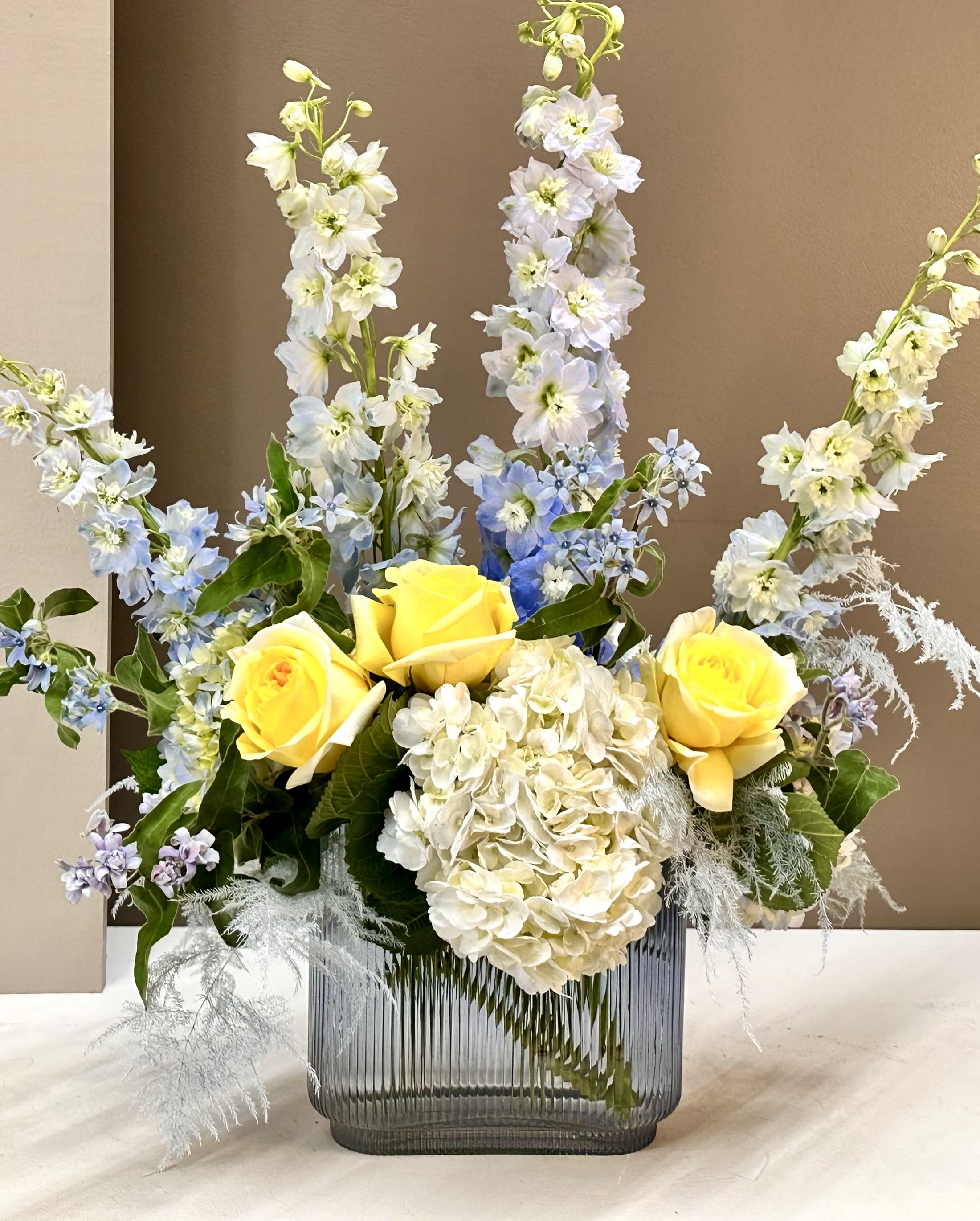Sunny Skies - A fresh, airy composition featuring sunny Yellow Roses, soft Blue Delphinium, and lush Hydrangea. Accented with delicate textures, this arrangement is styled in a modern glass vase for a light, elegant look. Perfect for brightening any space.