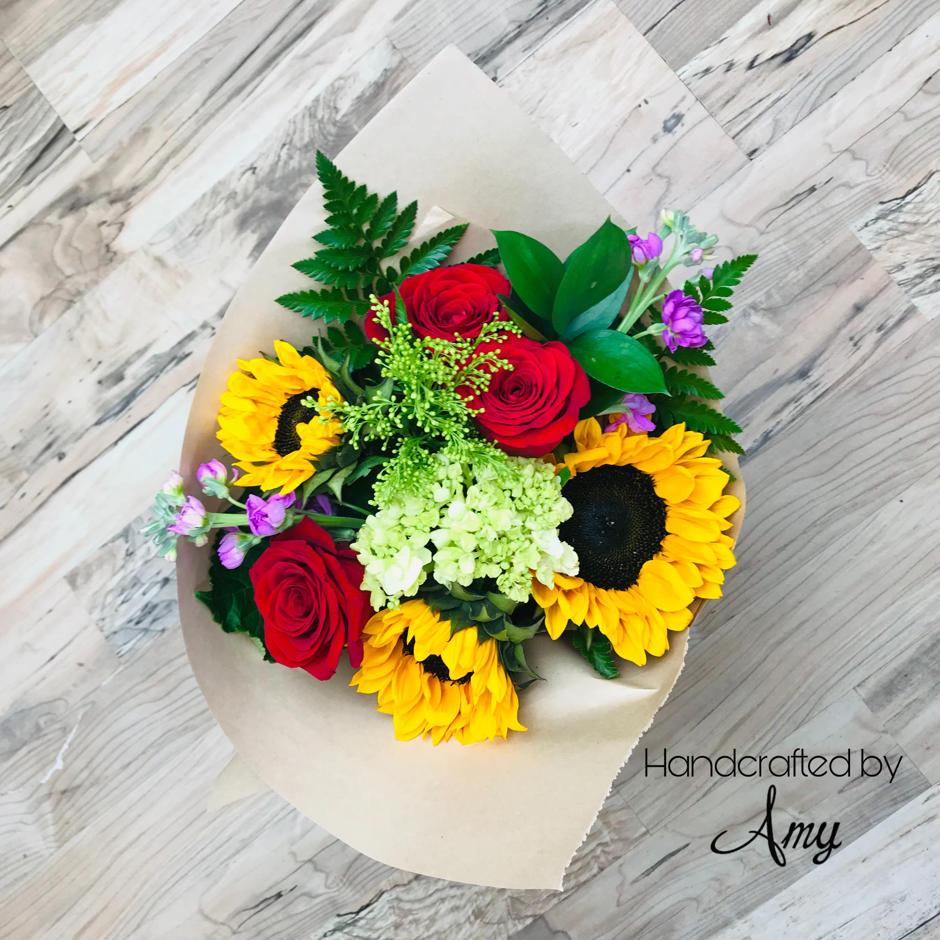 Wrapped bouquet of yellow sunflowers, red roses, hydrangea, and purple blooms on a wood background