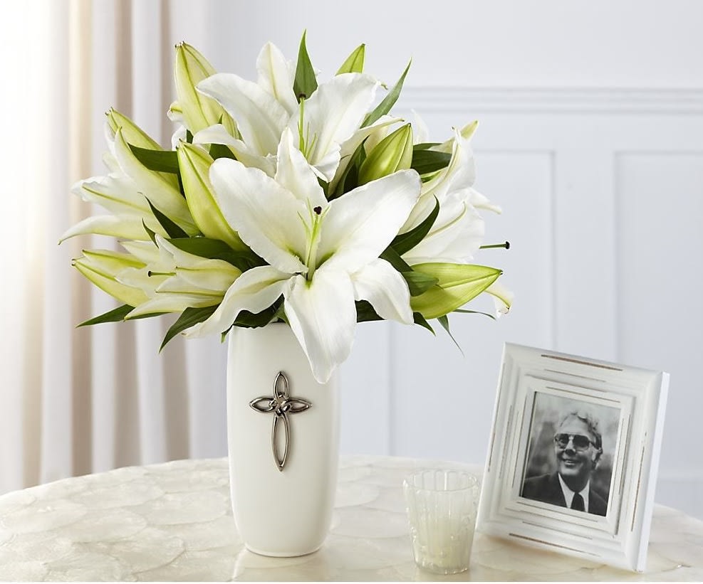 White lilies arranged in a tall white vase beside a framed photo