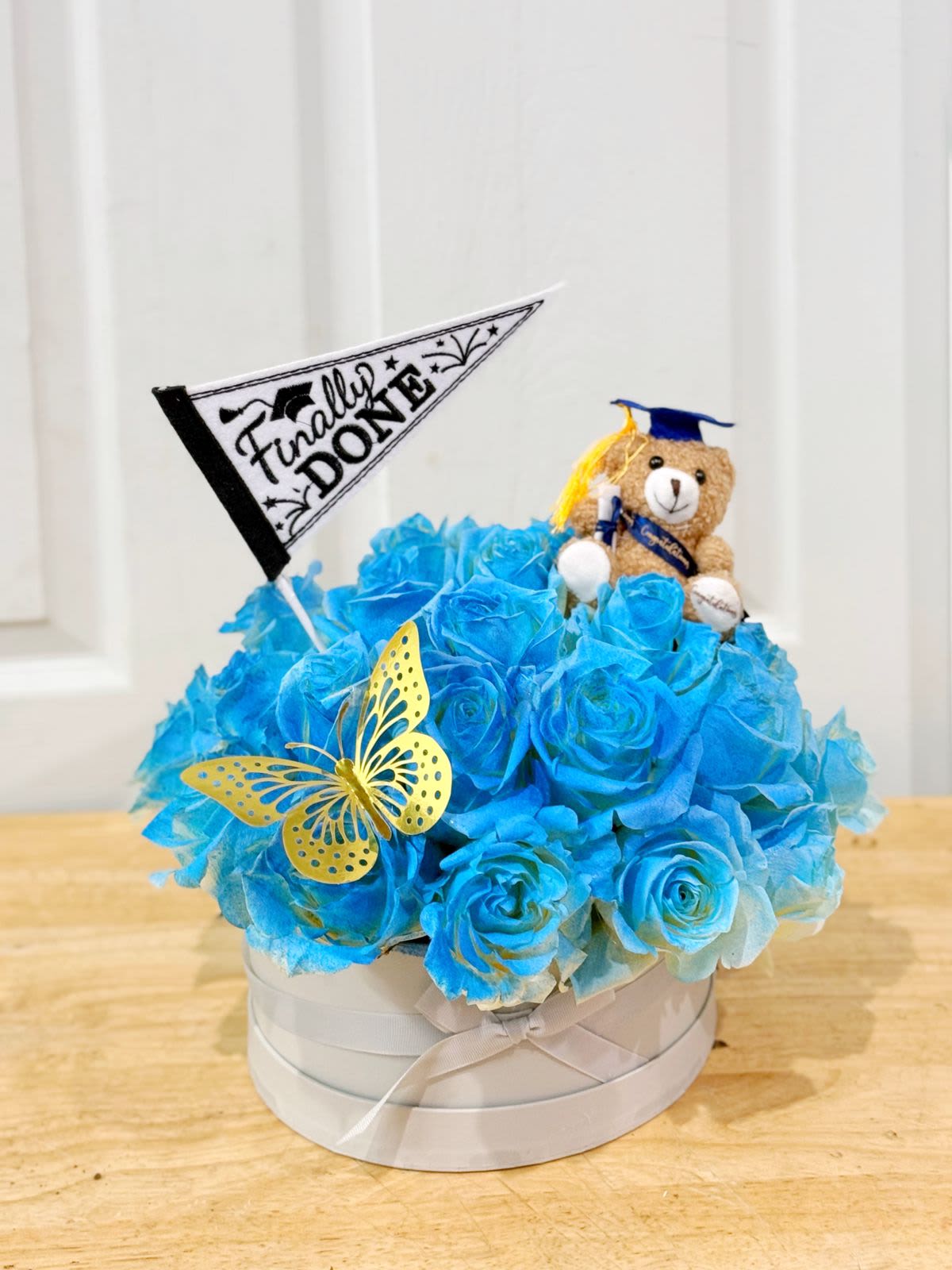 Blue rose arrangement in a round box with a teddy bear and graduation flag