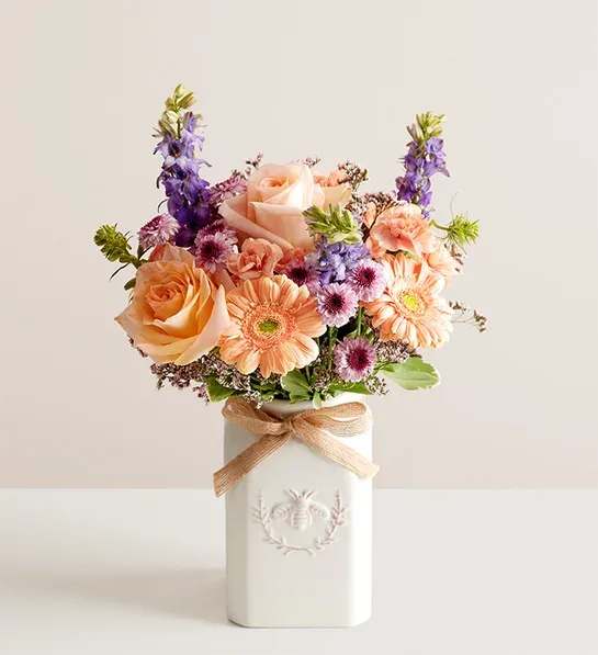 Honey Bee Buzz - Get them buzzing with our charming bouquet! A garden-fresh gathering of blooms in sweet shades of lavender, purple, peach and pink come together for a mix of color and texture. Designed in our Sweet As Can Bee artisanal vase, featuring an adorable honey bee and raffia bow, it’s a gift that adds quaint beauty to any decor.