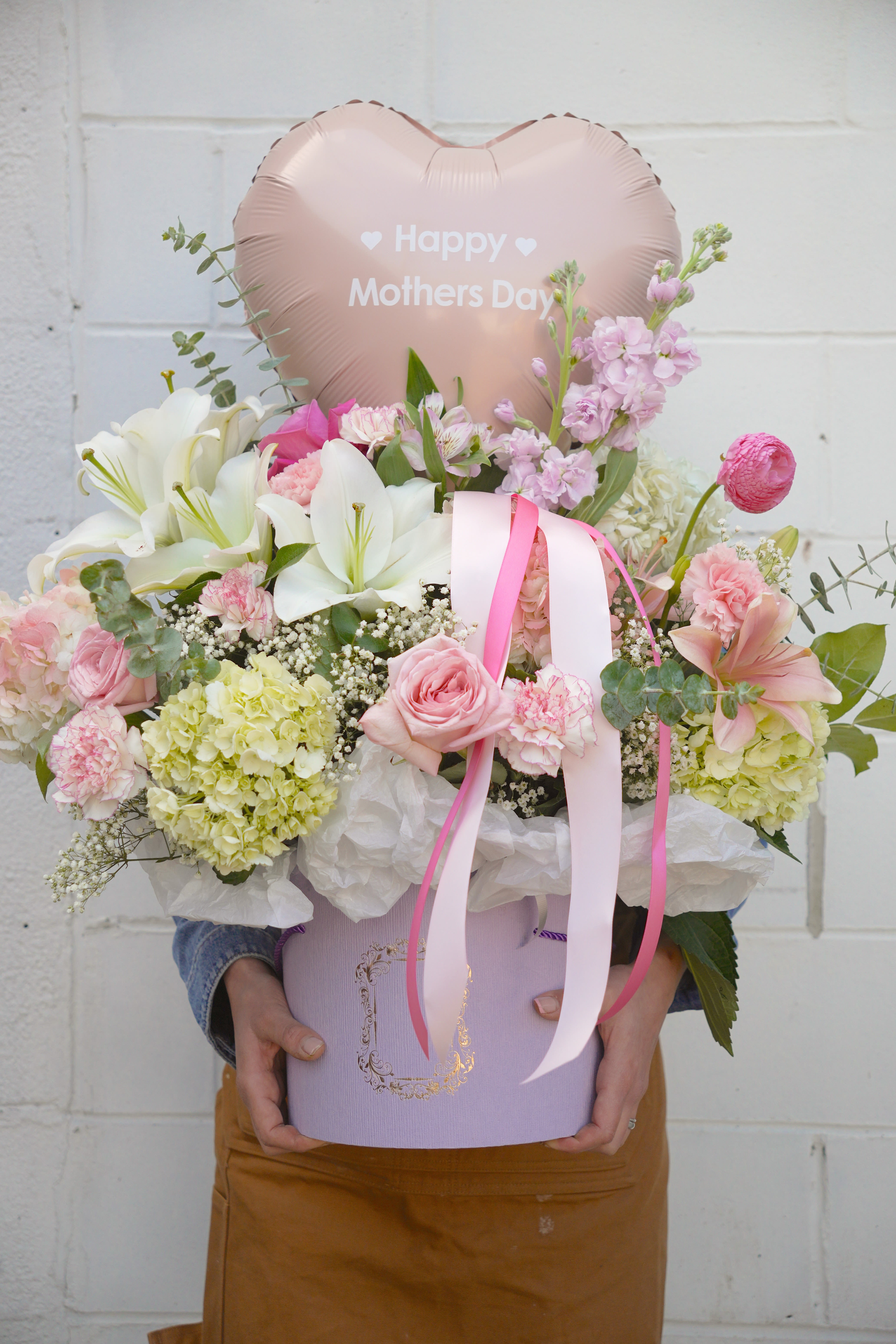 Mom's Sweet Love - Soft blush and white lilies and roses take center stage in this romantic Mother's Day bouquet, the kind of arrangement that makes Mom feel truly seen. Designed as a gift-ready presentation, the rounded hat-box style bouquet and large hand-held option both pair fragrant white lilies with pink roses, pale lavender hydrangeas, and soft carnations for a full, feminine look-finished with trailing pink and white ribbons and a pale pink heart-shaped "Happy Mothers Day" balloon for a celebratory touch.  The palette blends blush pink, white, pale lavender and fresh green accents for a layered, textured effect: large lilies and roses, fluffy hydrangea clusters, ruffled carnations, alstroemeria and delicate filler blooms create depth and fragrance. Each arrangement arrives in a lavender hat box or gift box with decorative tissue paper and a coordinating ribbon, presented polished and ready to place on a table or bedside. 