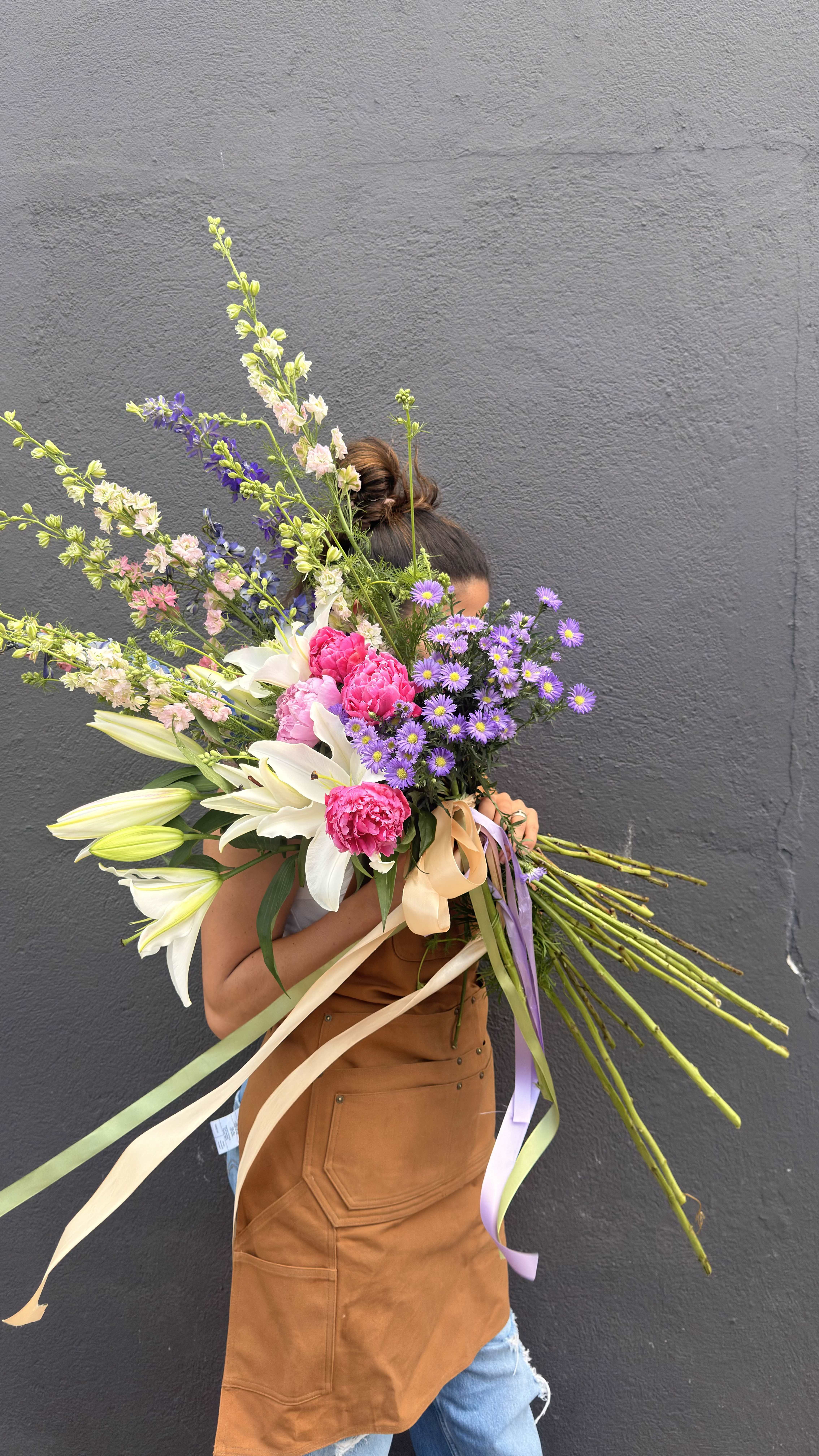 WILDFLOWER SERENADE - A lush hand-tied bouquet that reads like a sunlit meadow-perfect as a birthday bouquet, anniversary arrangement, or a romantic wedding gift. White lilies and bright pink peonies take center stage, while sprays of small purple daisy-like blooms and snapdragons add a playful, garden-fresh texture that makes the recipient feel cherished and celebrated.