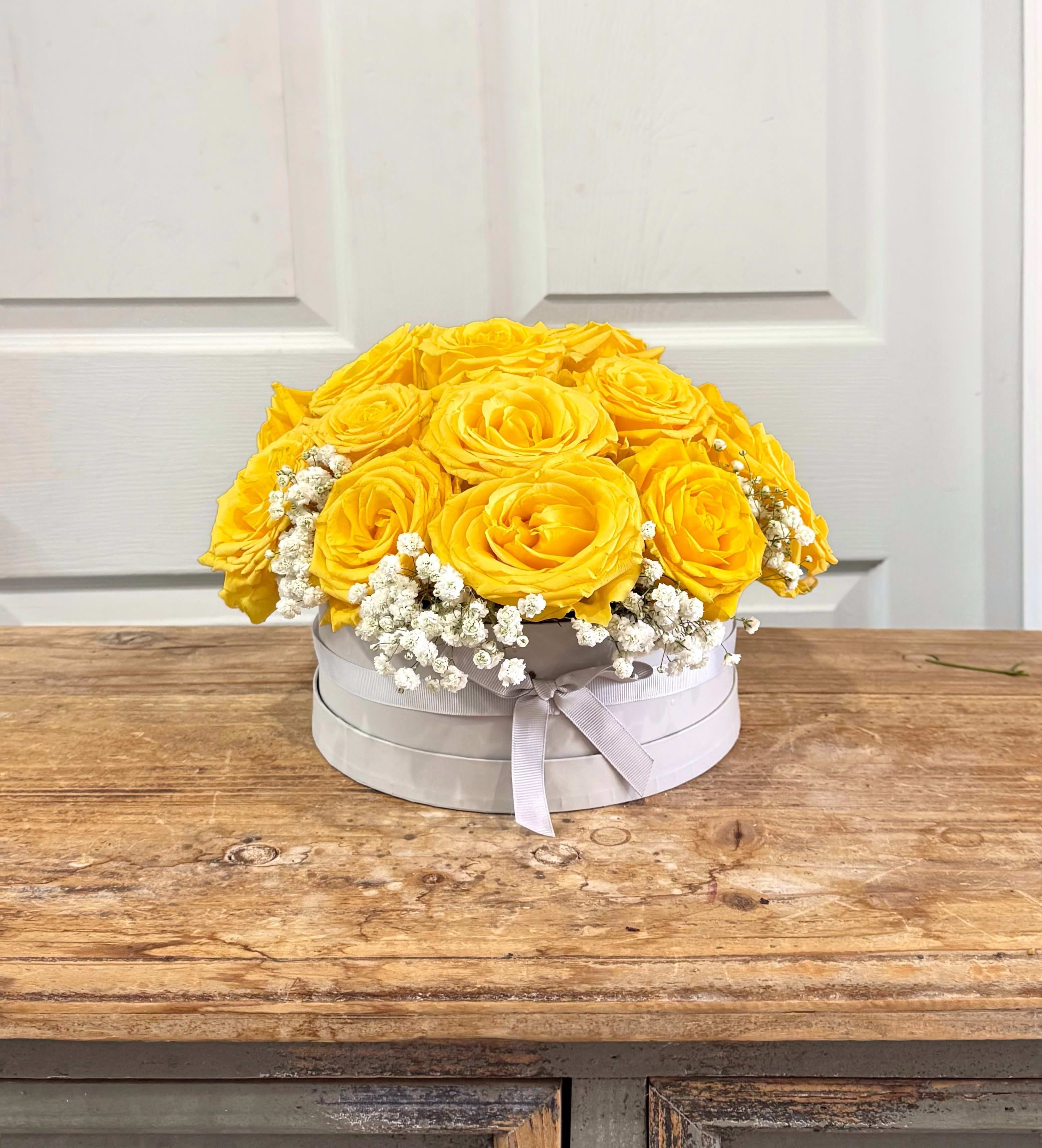 Round white box of yellow roses with small white filler flowers on a wooden table