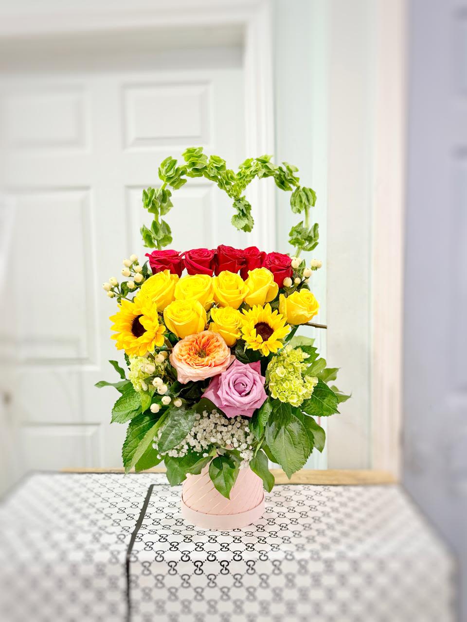 Heart of Joy - A colorful way to say “I care.” Our Heart of Joy arrangement celebrates love, happiness, and gratitude with a vibrant combination of blooms. Featuring a joyful mix of red and yellow roses, sunflowers, lavender and peach roses, hydrangea, and Bells of Ireland shaped into a delicate heart, this arrangement is pure visual poetry.  Carefully arranged in a soft pink ceramic vase, this design brings together elegance and playfulness, making it perfect for birthdays, anniversaries, or simply brightening someone’s day.  A little note for you, who choose with love: All our arrangements are handmade with fresh flowers that bloom naturally. That means some flowers may be more open or closed compared to the photo. Also, depending on the season and store availability, certain shades or flower types may vary slightly. If any flowers are unavailable, thoughtful substitutions will be made to preserve the overall look, feel, and beauty of your arrangement.