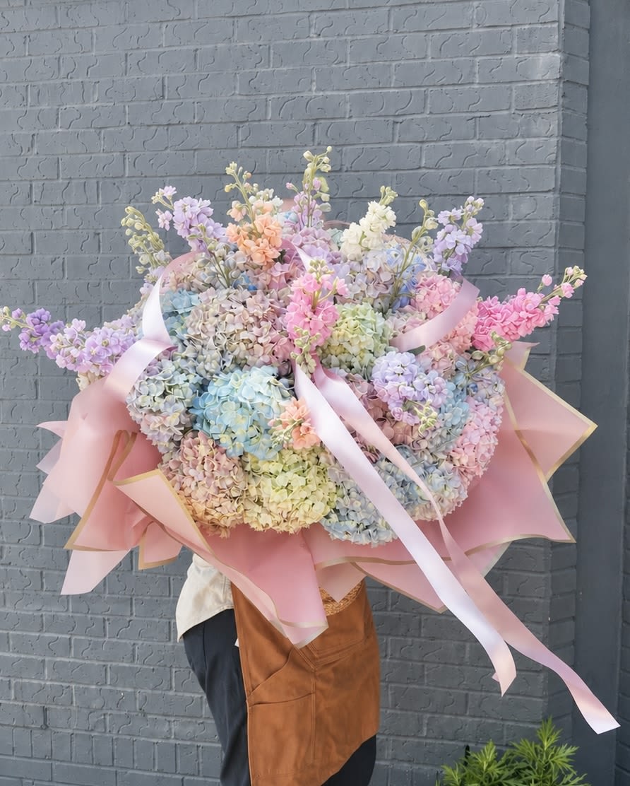 COTTON CANDY  DREAMS - An oversized, hand-held pastel bouquet that reads like a daydream-perfect as a wedding bouquet, birthday surprise, or spring celebration. Cotton Candy Dreams uses large, rounded masses of hydrangeas in soft pink, lavender, pale blue, cream and pale yellow to create an immediate visual impact; tall stock stems rise above the blooms for airy height and a touch of graceful movement. The overall impression is romantic and celebratory-the kind of arrangement that makes the person you're thinking of feel truly seen.