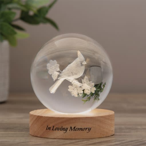 Crystal Keepsake Cardinal Orb With Wood LED Base - A delicate glass orb featuring a white cardinal and tiny white floral accents offers a quiet, heartfelt way to say you're thinking of someone - a sympathy arrangement and memorial keepsake meant to bring comfort. The rounded wooden LED base engraved with "In Loving Memory" casts a gentle glow through the transparent sphere, creating a soft, contemplative presence that suits remembrance moments and condolence gifting.  Inside the clear glass, the sculpted white bird and miniature blossoms float against the open space of the orb, their restrained white-and-natural palette emphasizing peace and simplicity. The warm natural wood tone of the base grounds the piece, while the subtle LED illumination highlights the delicate details without overpowering them. Compact and tasteful, this keepsake fits on a mantel, bedside table, or memorial display.  Offer this to someone grieving or honoring a loved one as a lasting token of remembrance; it's the kind of gift that helps a person feel seen and comforted. The engraved base and included LED light make it an easy-care, enduring memento - no watering needed, just place it where its soft glow can provide quiet solace.