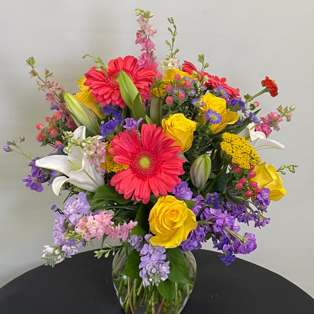 Colorful mixed bouquet in a clear glass vase with gerbera daisies, roses, lilies, and purple filler flowers