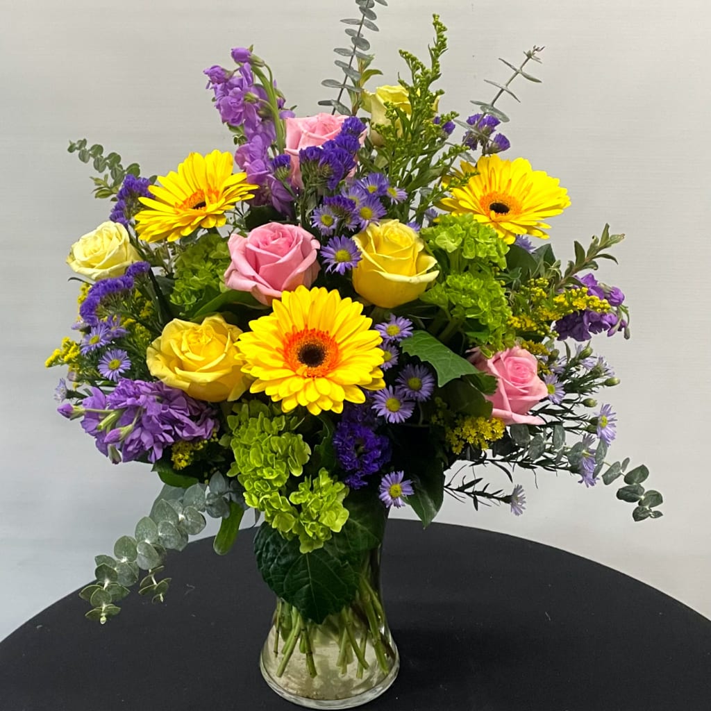 Mixed bouquet of yellow gerbera daisies, pink and yellow roses, and purple flowers in a glass vase