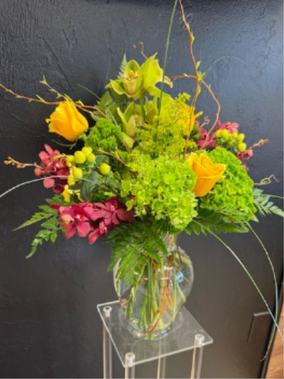 Tall arrangement of orange roses, green hydrangea, and orchids in a clear glass vase