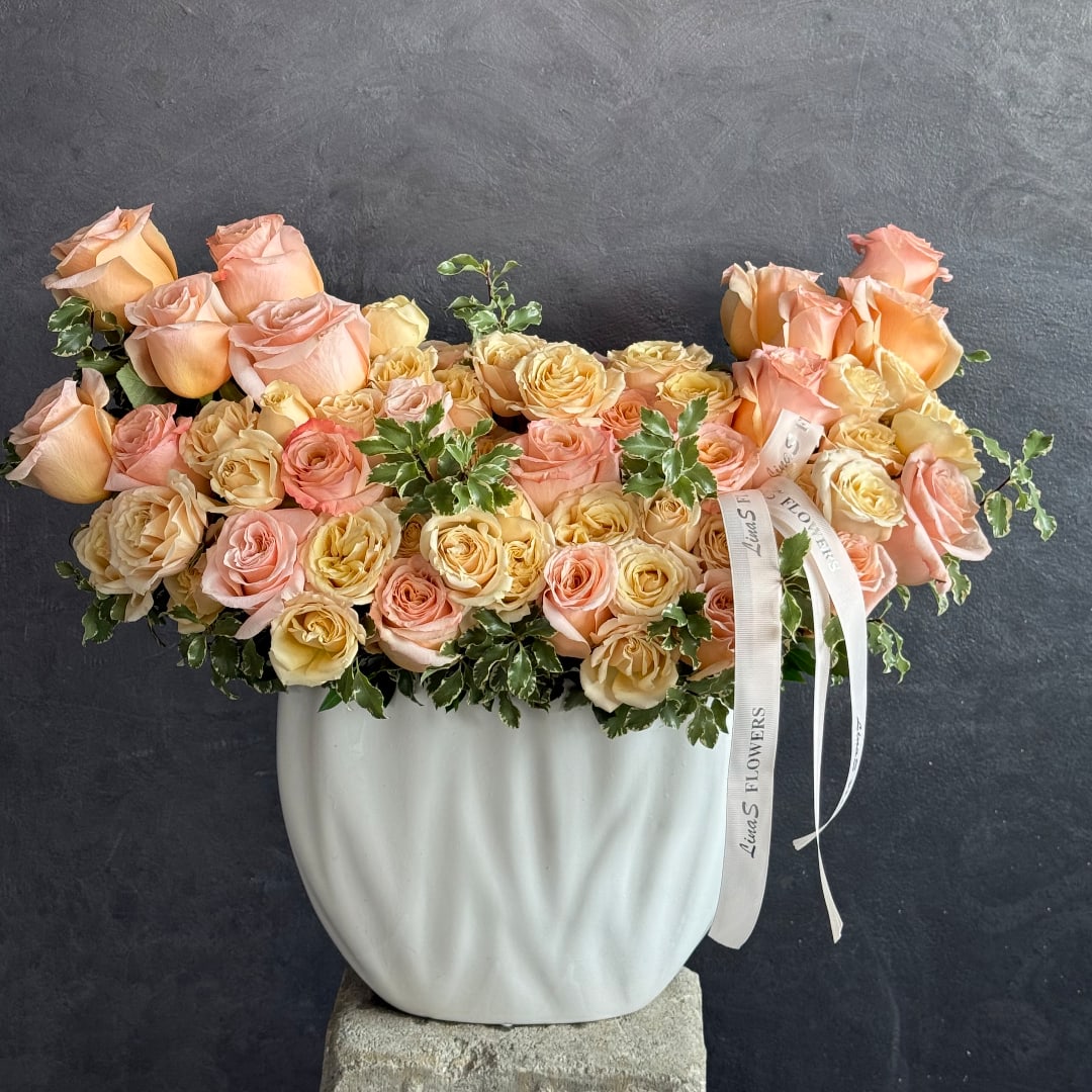 Peach and blush roses arranged in a white vase with ribbon accents