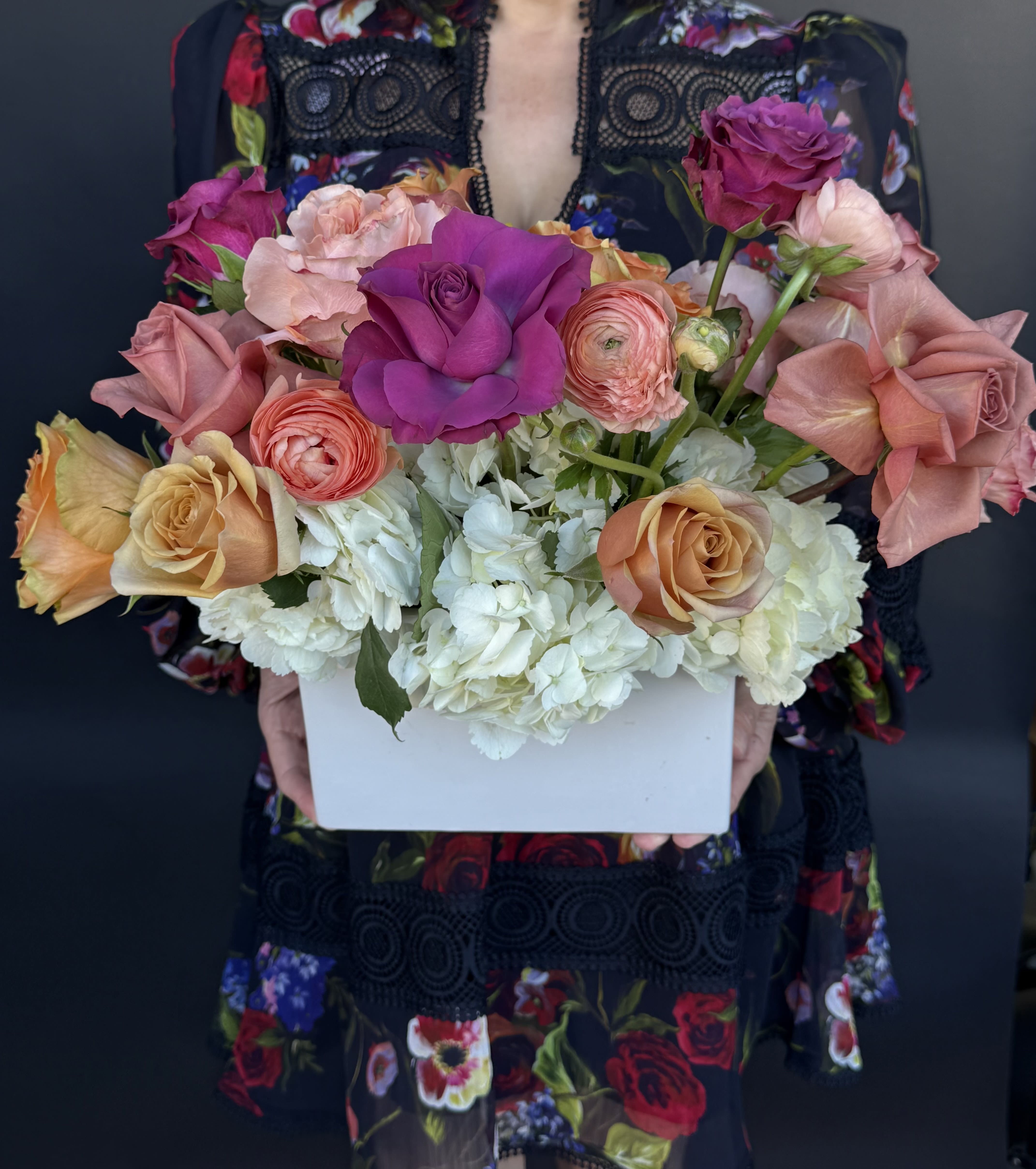 Colorful arrangement of roses and ranunculus with white hydrangeas in a white rectangular box.