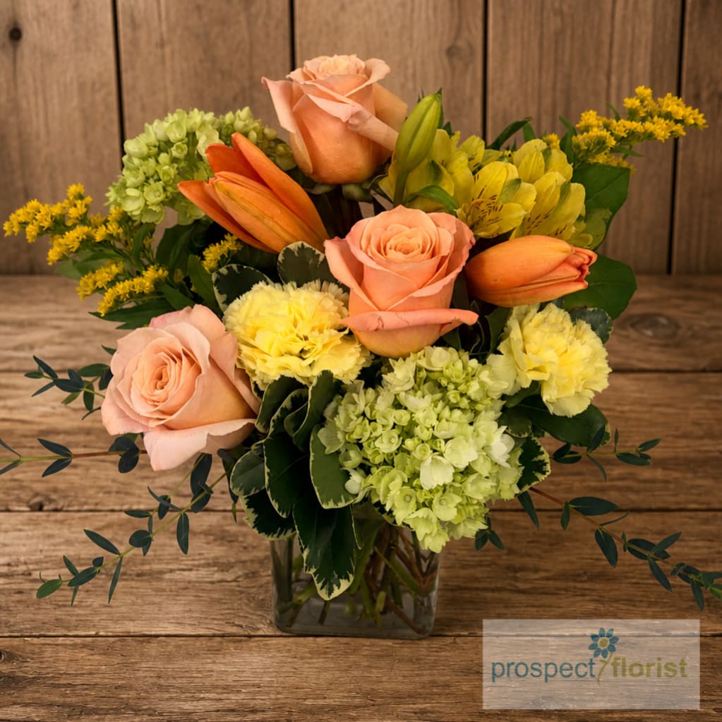 Mixed bouquet of peach roses, lilies, and yellow flowers in a glass vase