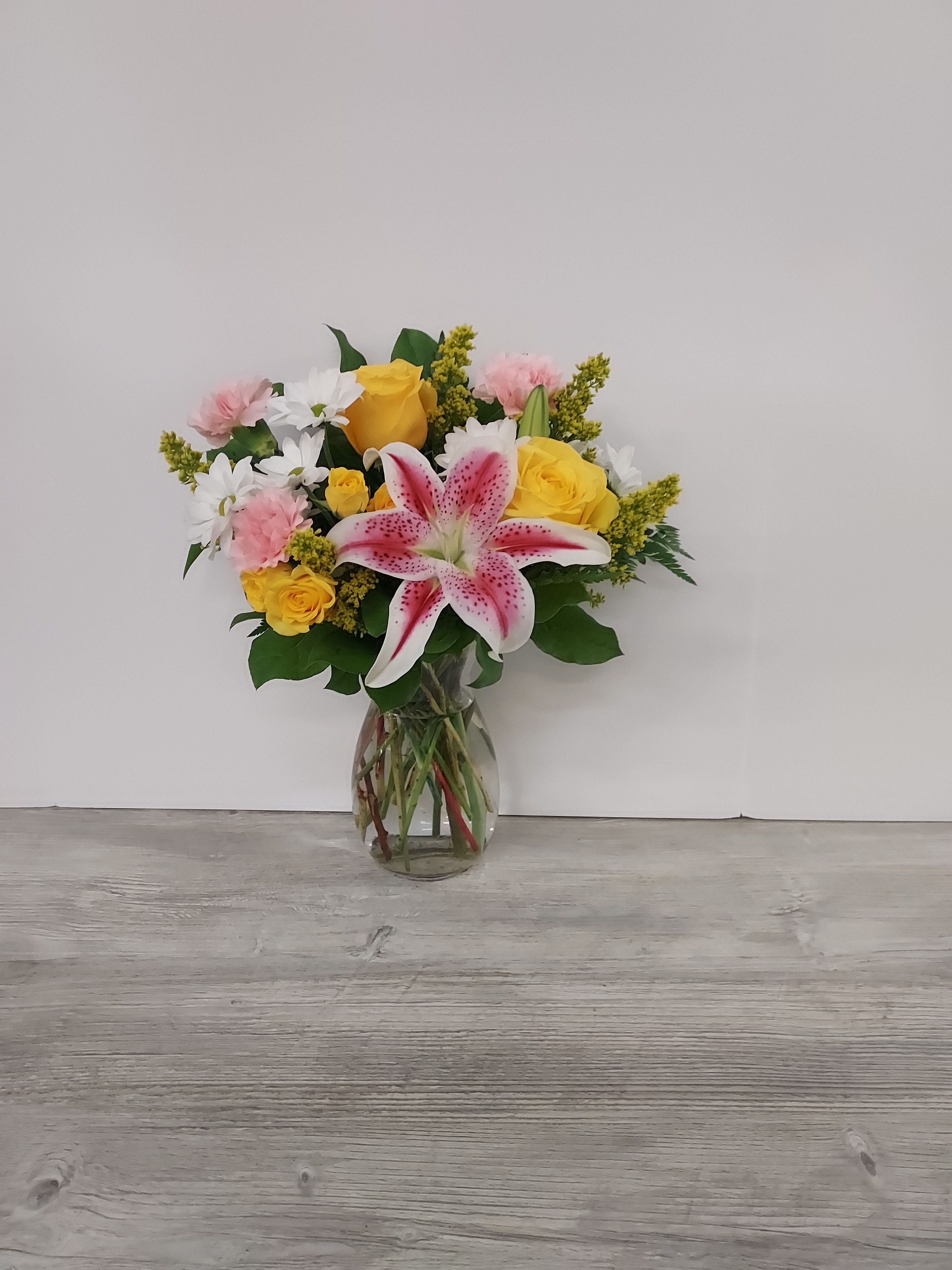 Mixed bouquet of pink lilies, yellow roses, white daisies, and carnations in a clear glass vase