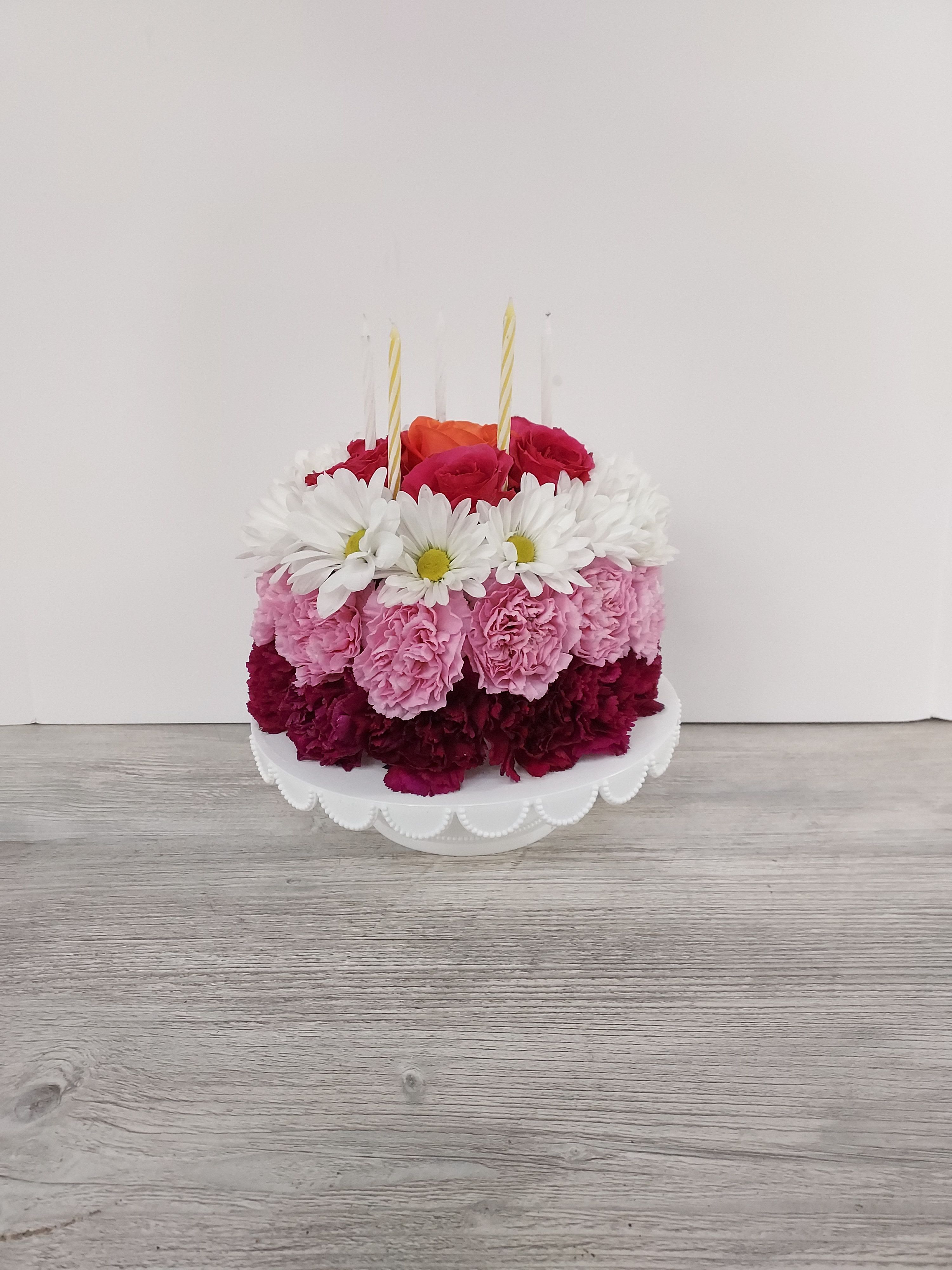 Round floral cake of pink carnations, white daisies, and roses with candles on a white cake stand