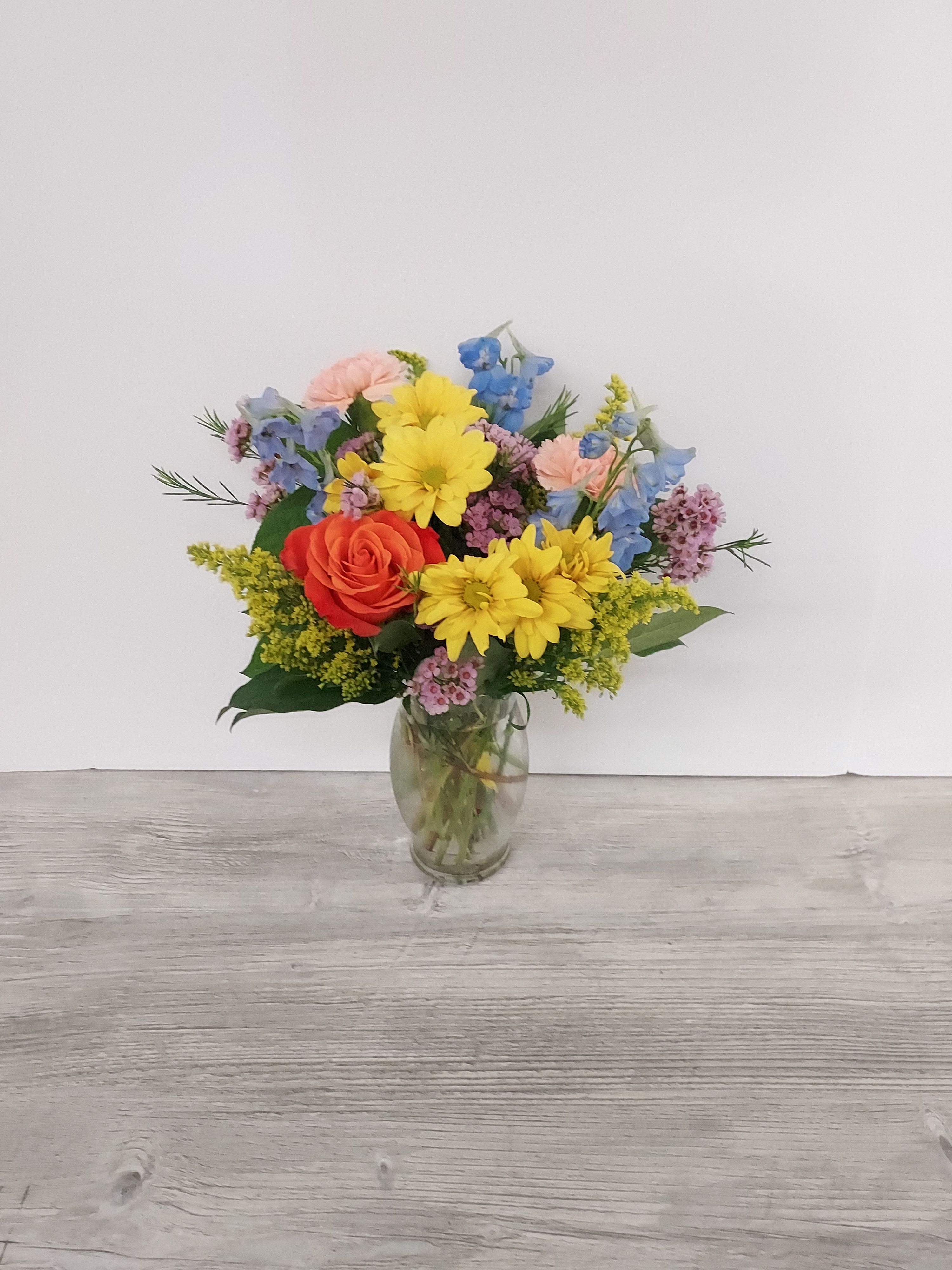 Mixed bouquet of yellow daisies, orange rose, and pastel flowers in a clear glass vase