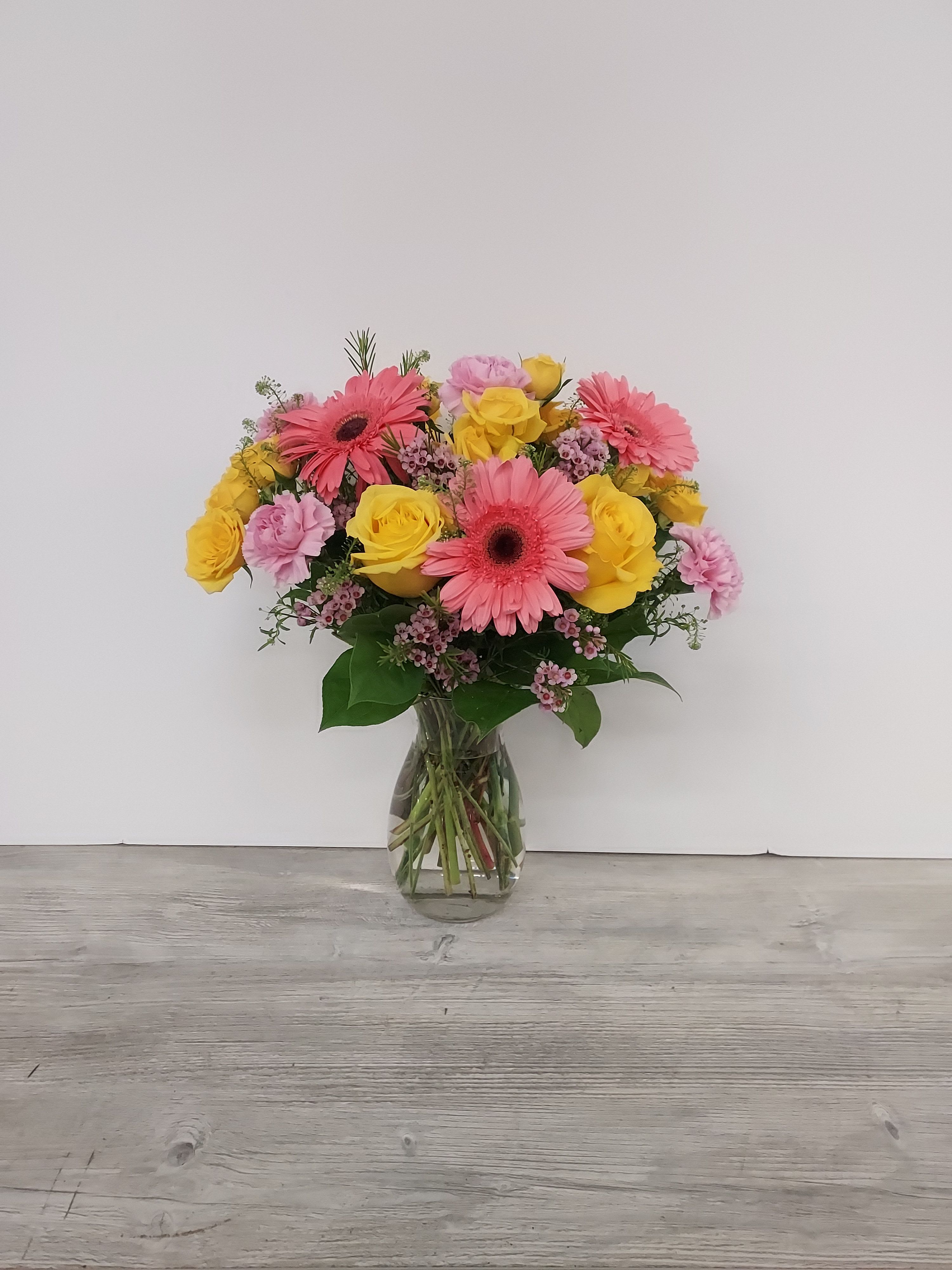 Pink gerbera daisies, yellow roses, and pink carnations arranged in a clear glass vase