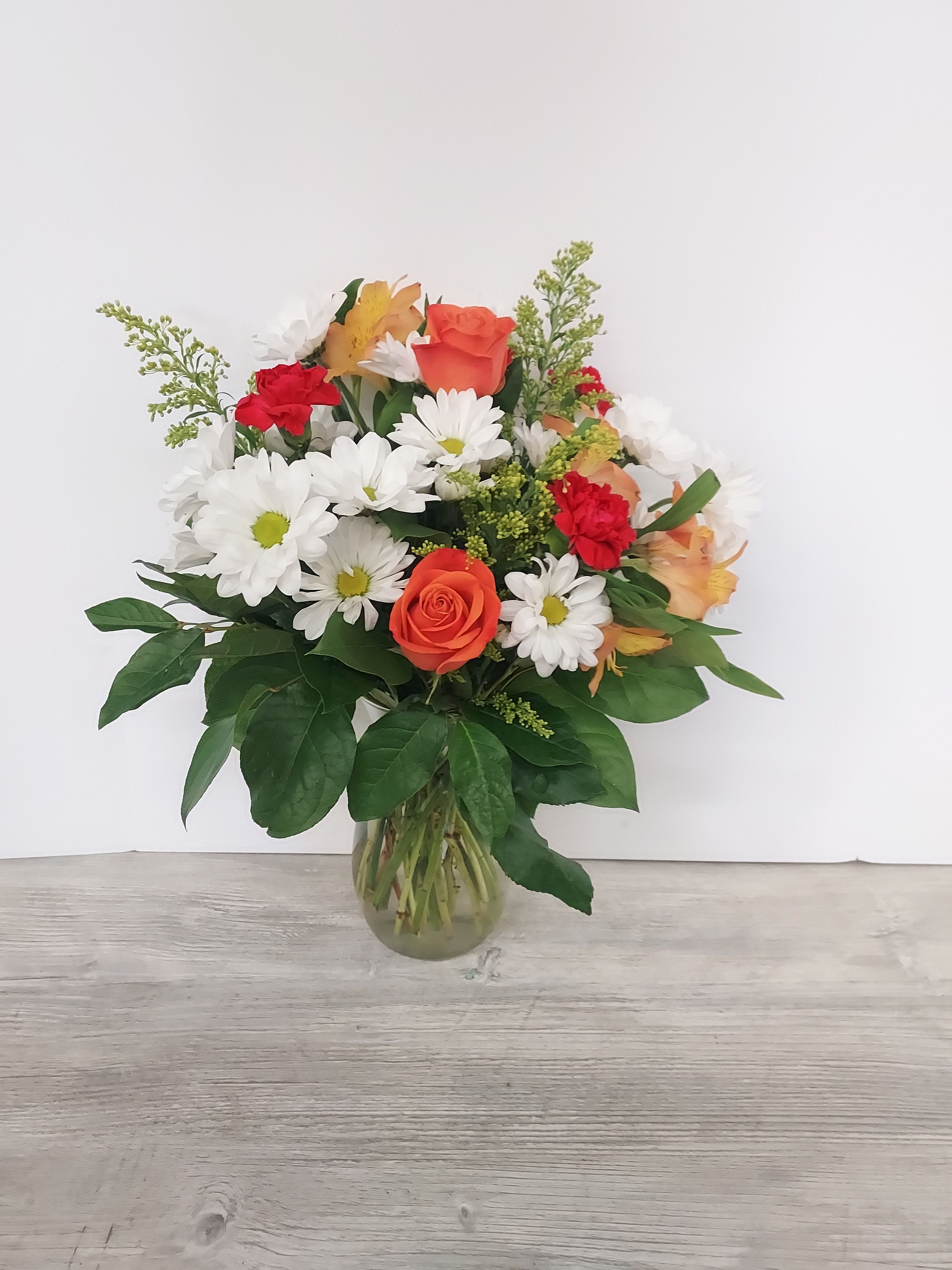 Mixed bouquet of orange roses, red carnations, and white daisies in a clear glass vase