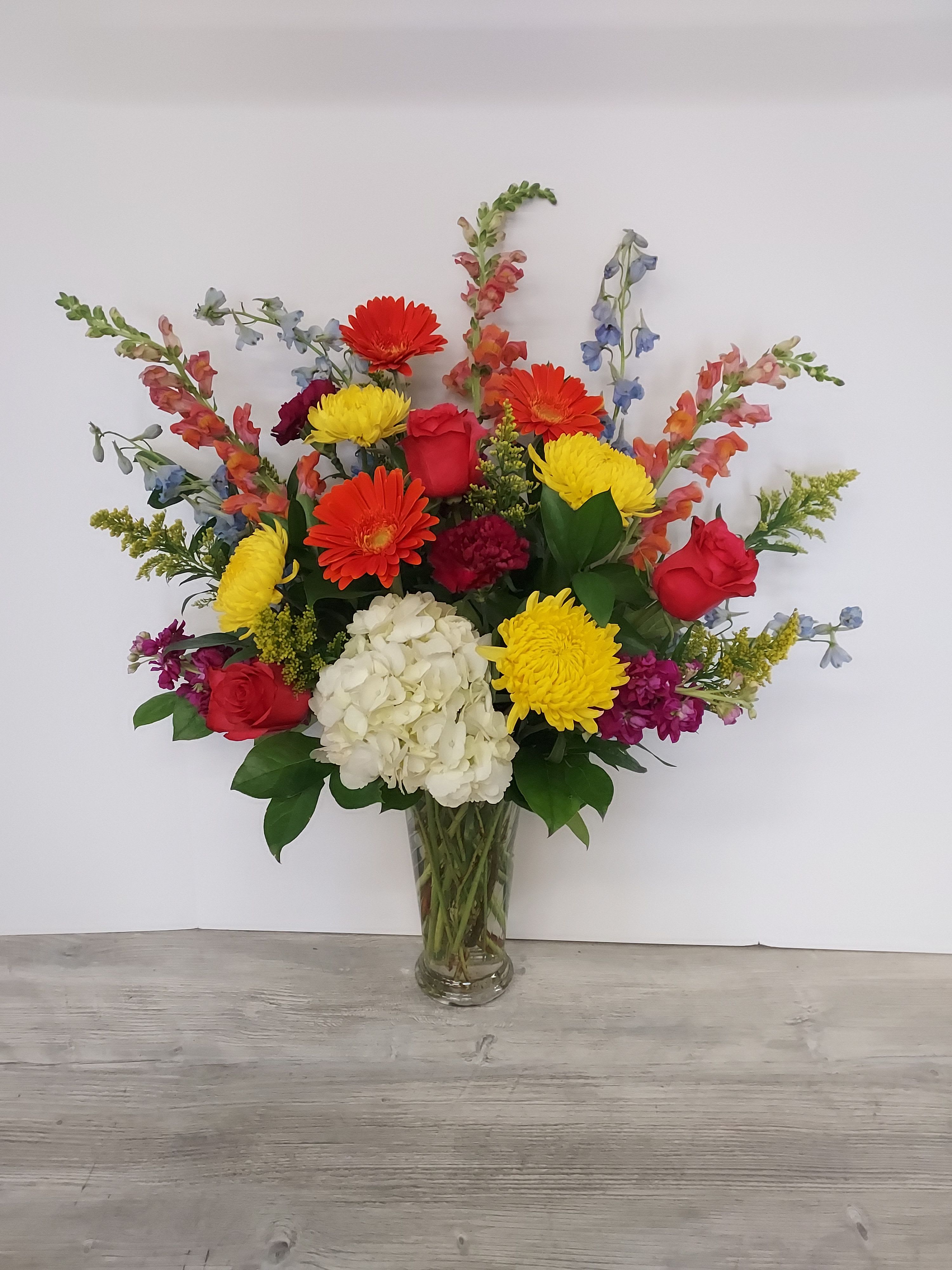 Tall mixed arrangement of red roses, orange gerbera daisies, yellow mums, and white hydrangea in a clear vase