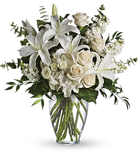 Tall arrangement of white lilies, roses, and other white flowers in a clear glass vase